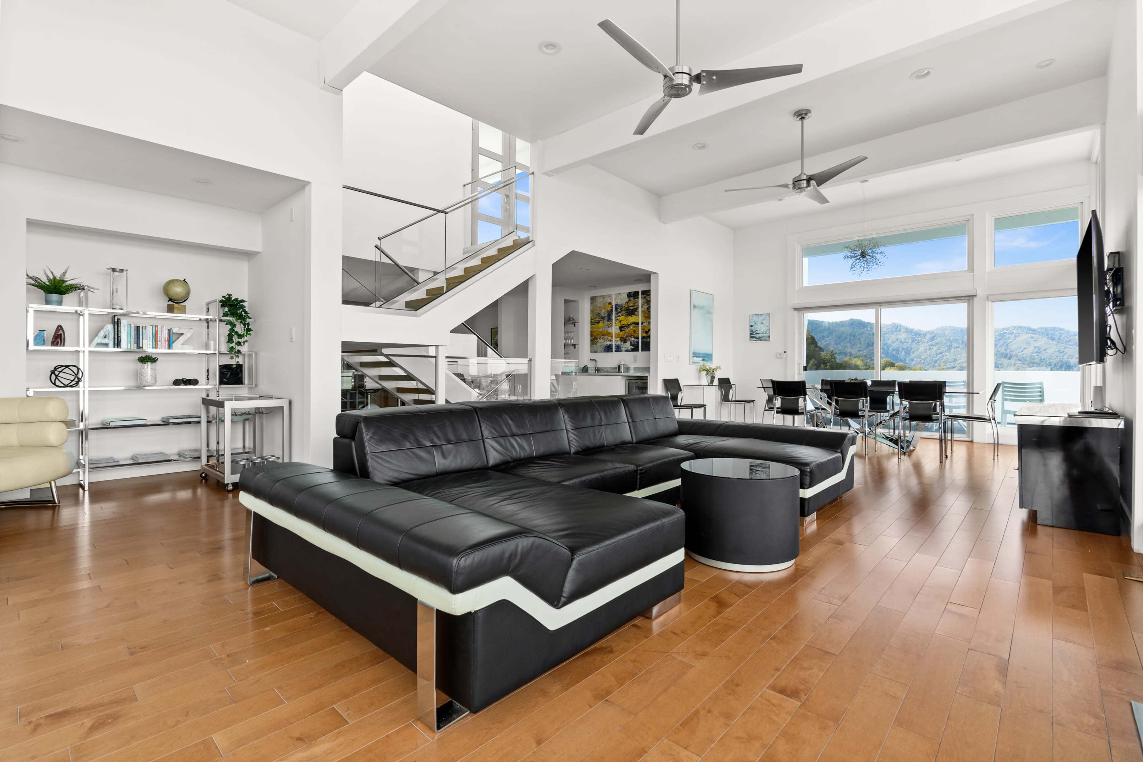 A modern living room features a large black sectional sofa, wooden flooring, and large windows that provide a view of the mountains.