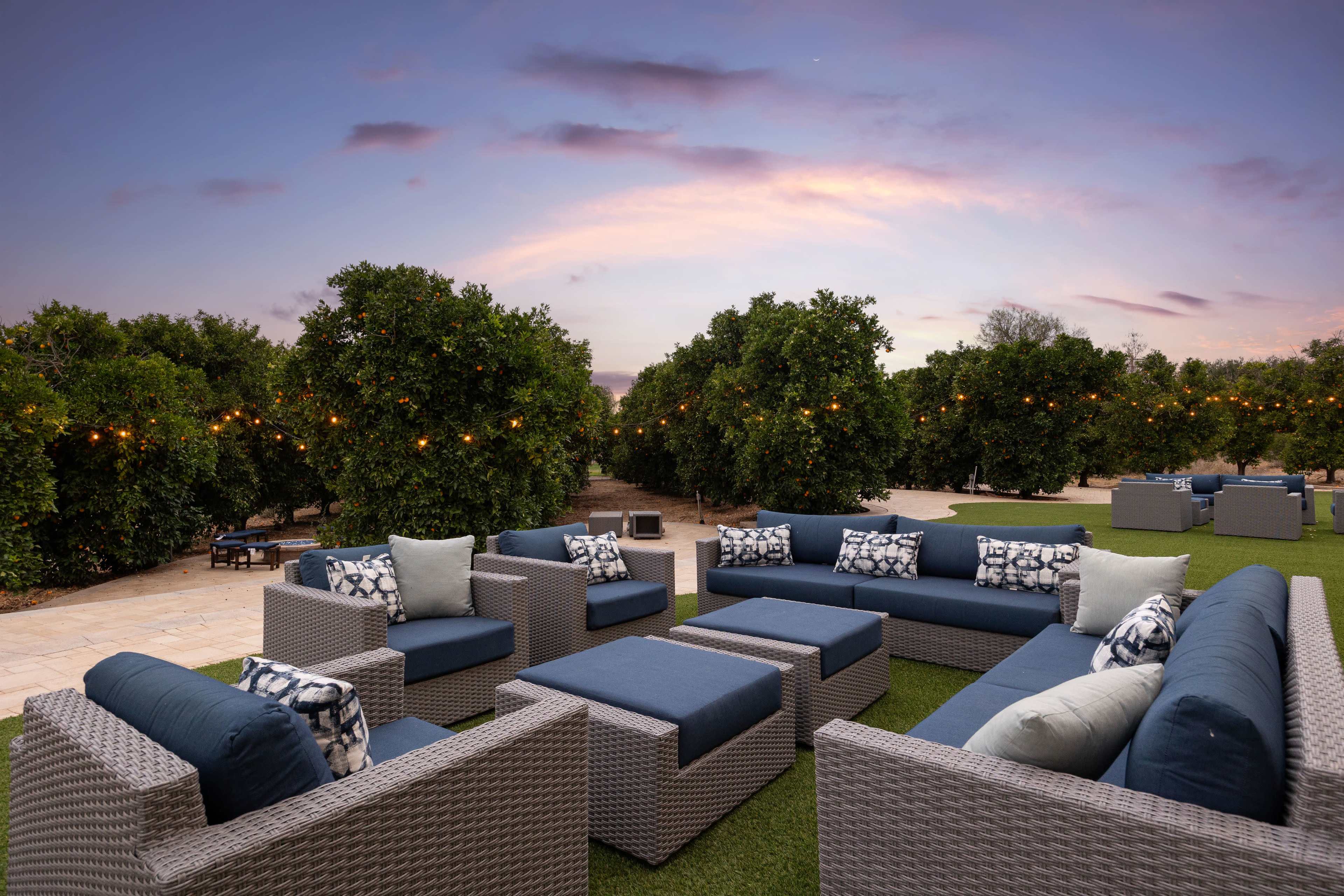 A spacious outdoor seating area features sectional sofas arranged around a central table, surrounded by lush greenery and soft evening lighting.