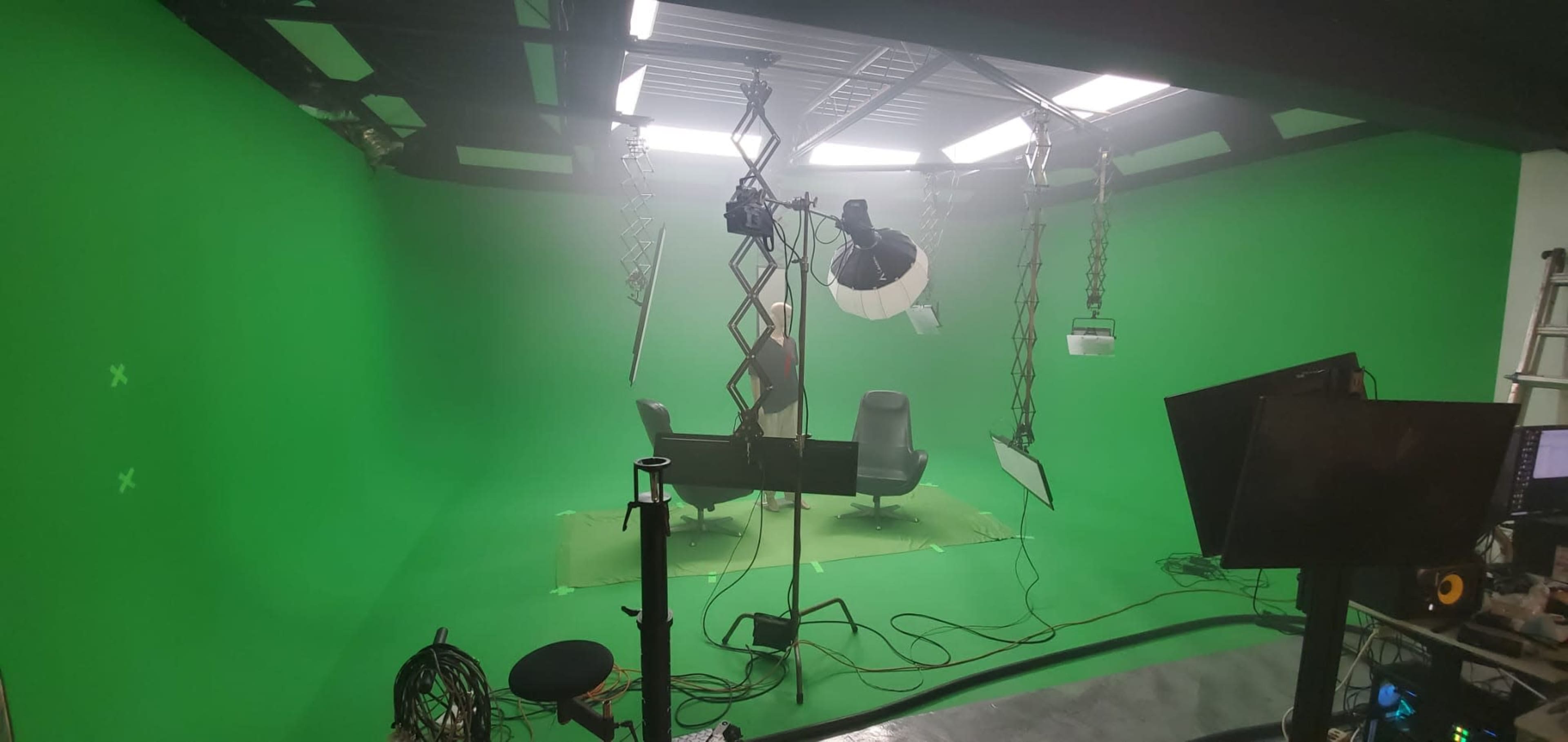 The image shows a studio setup with a green backdrop, lights, and two black chairs arranged around a table.