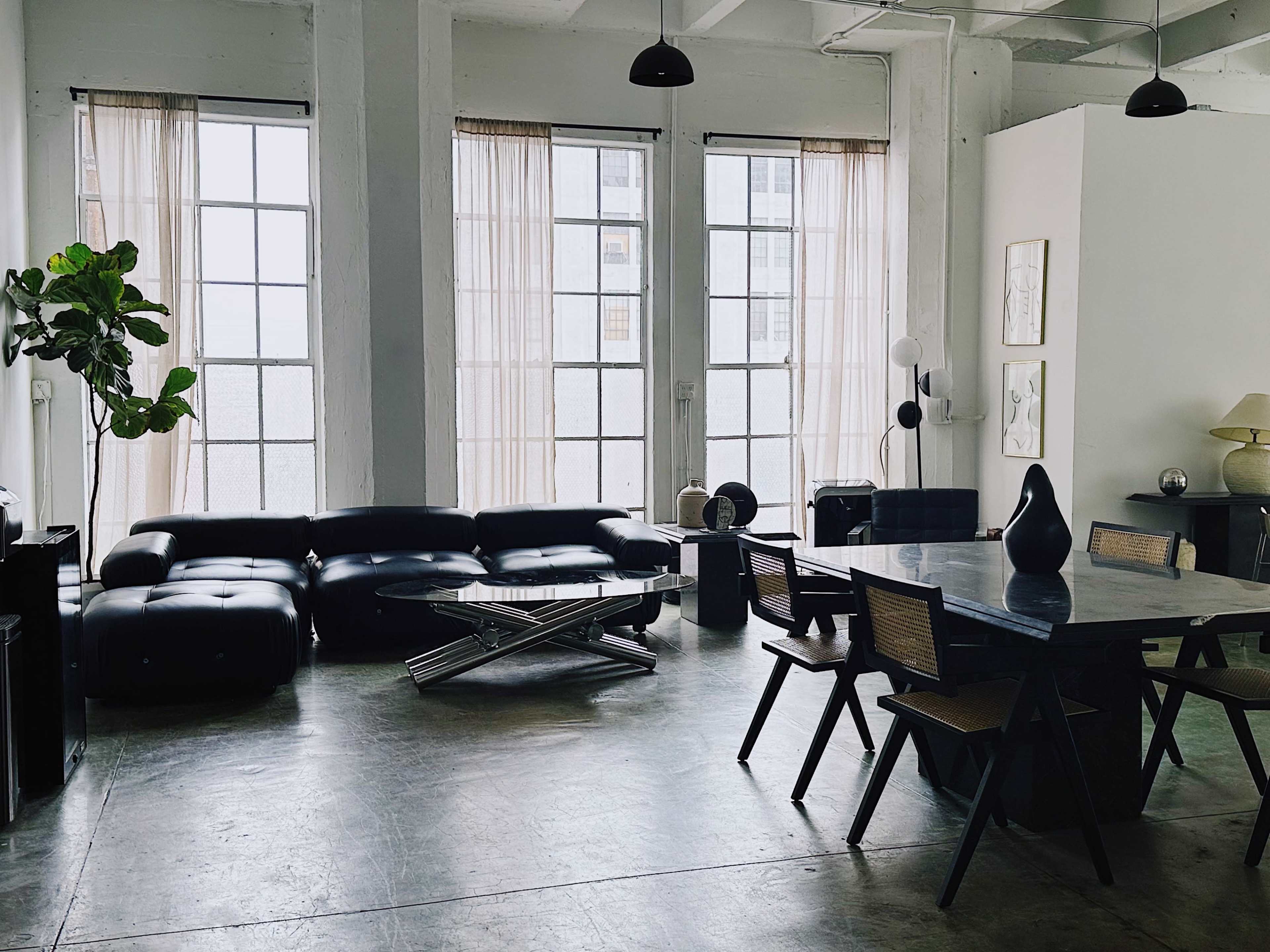 A spacious, industrial-style living area with large windows, a black sectional sofa, a modern dining table, and various decorative elements.