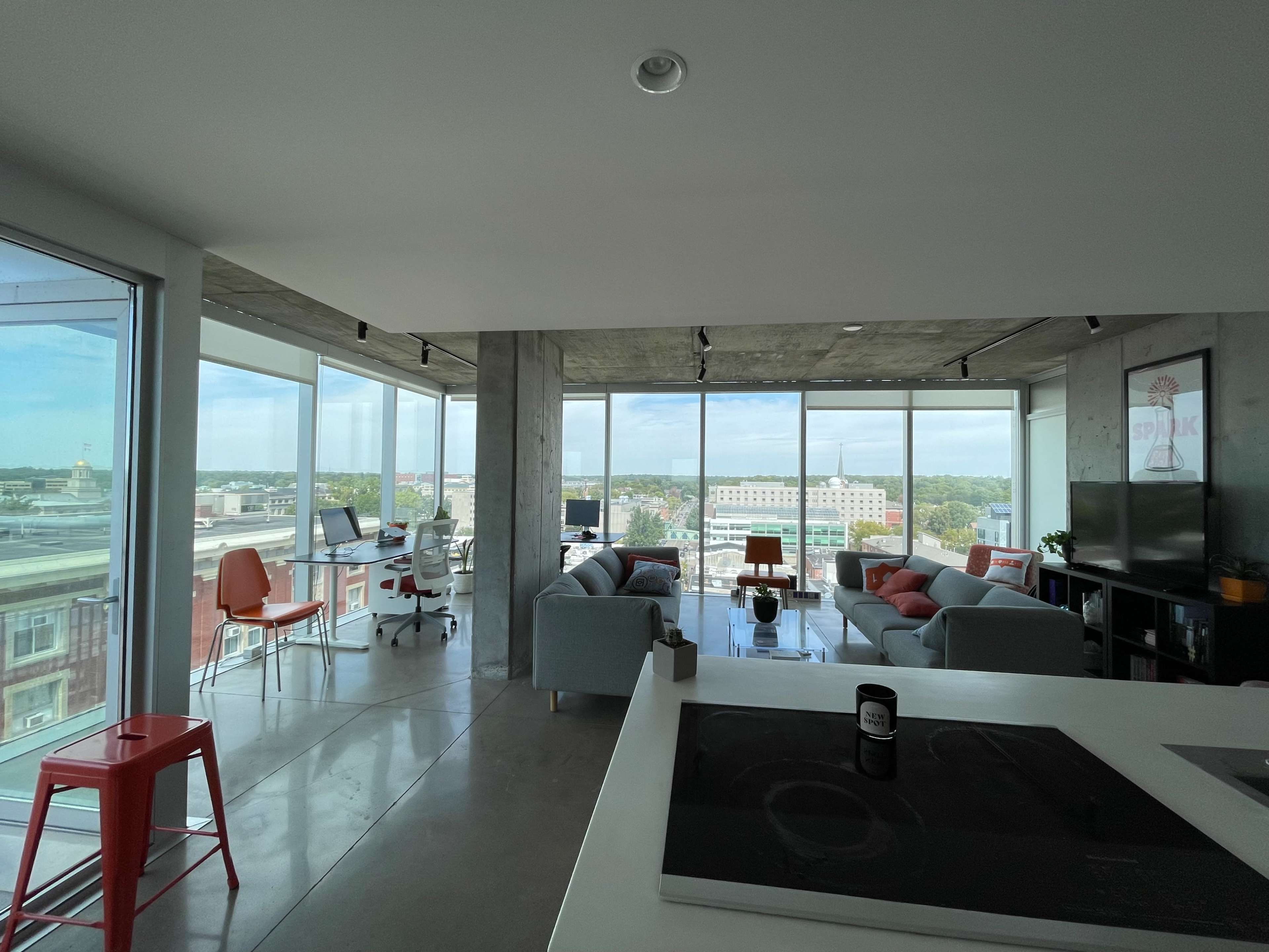 A modern open-concept living space with large windows, offering a view of the city and featuring a kitchen area, comfortable seating, and workstations.
