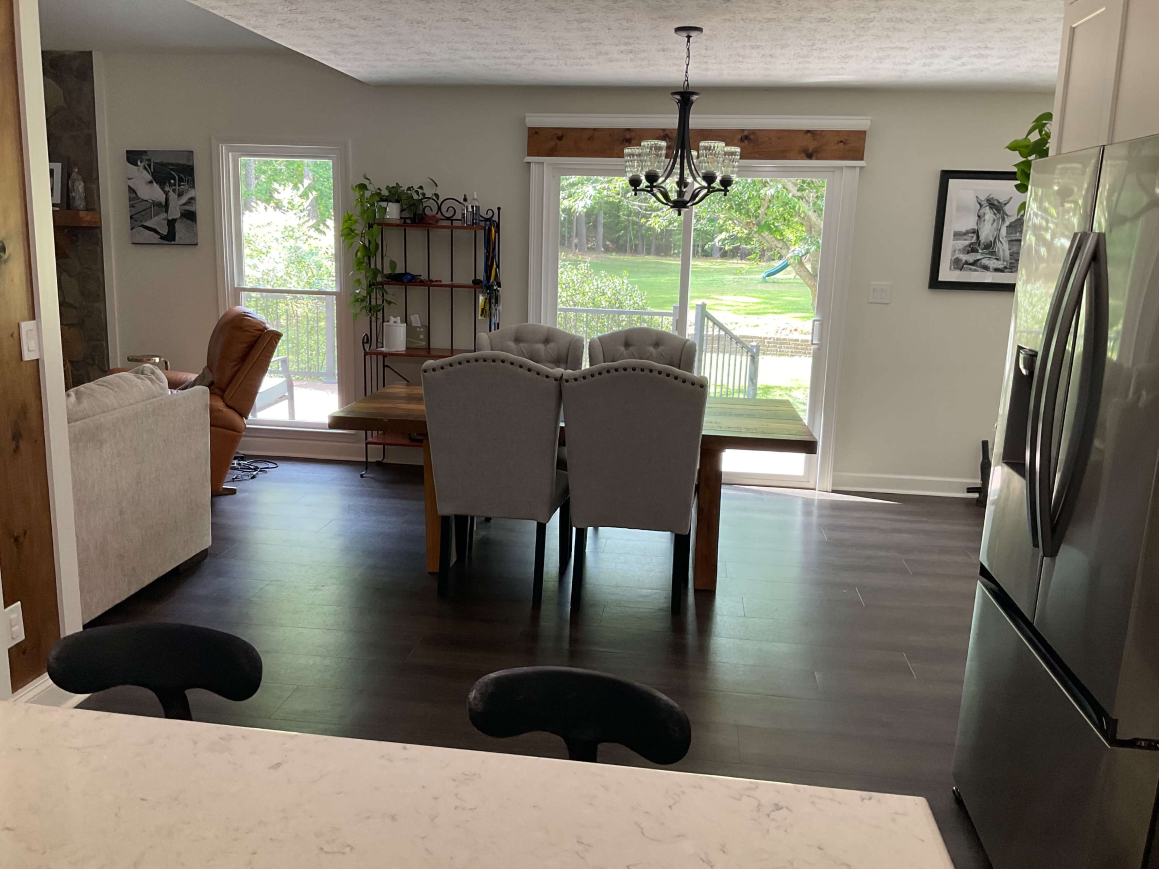 The image shows a dining area with a wooden table and gray upholstered chairs, adjacent to a living space featuring a brown recliner and large windows overlooking a yard.