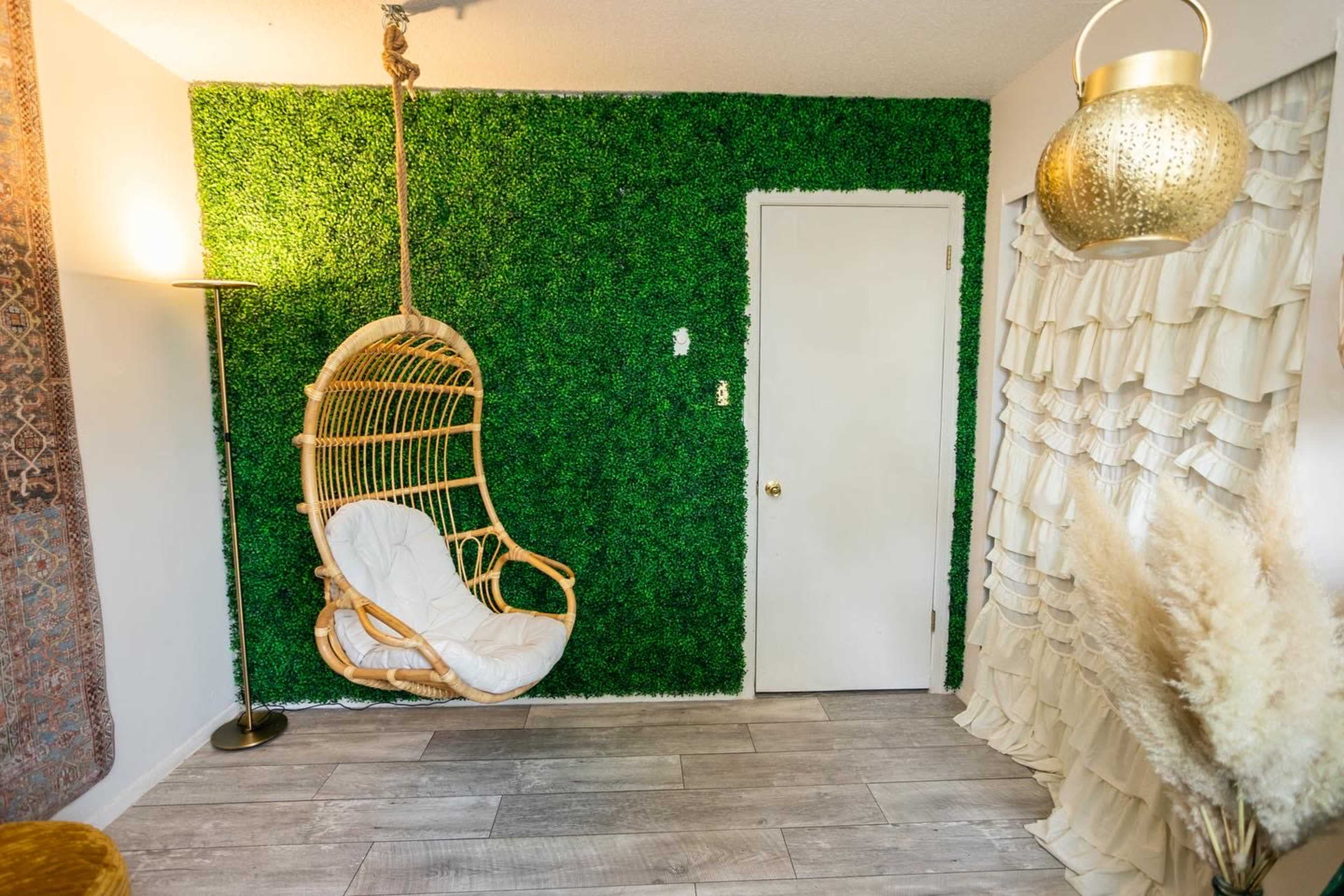A room featuring a hanging rattan chair against a green artificial grass wall, with a door and textured curtains nearby.