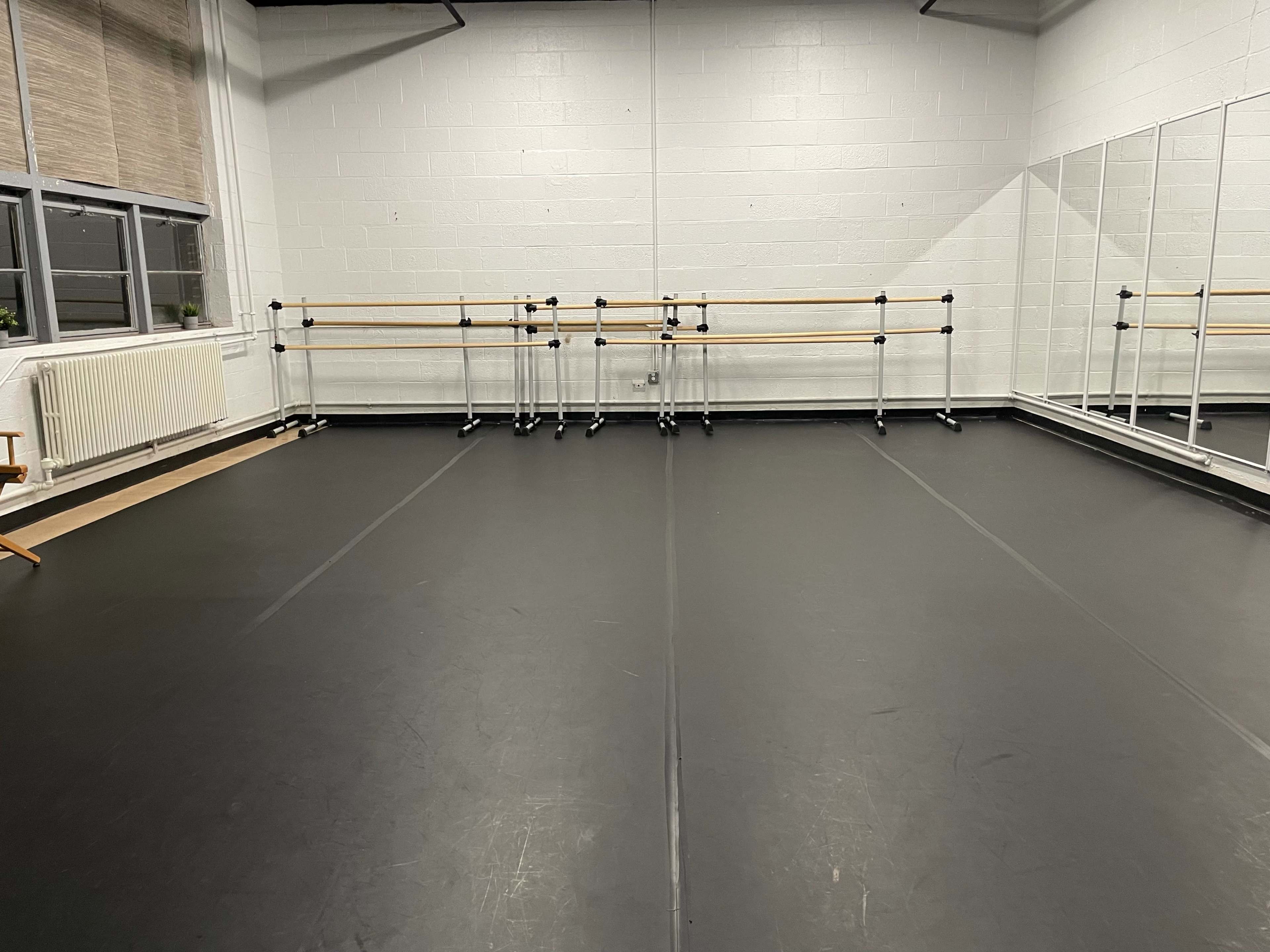 A spacious dance studio with a black floor, mirrored wall, and ballet bars positioned against the back wall.