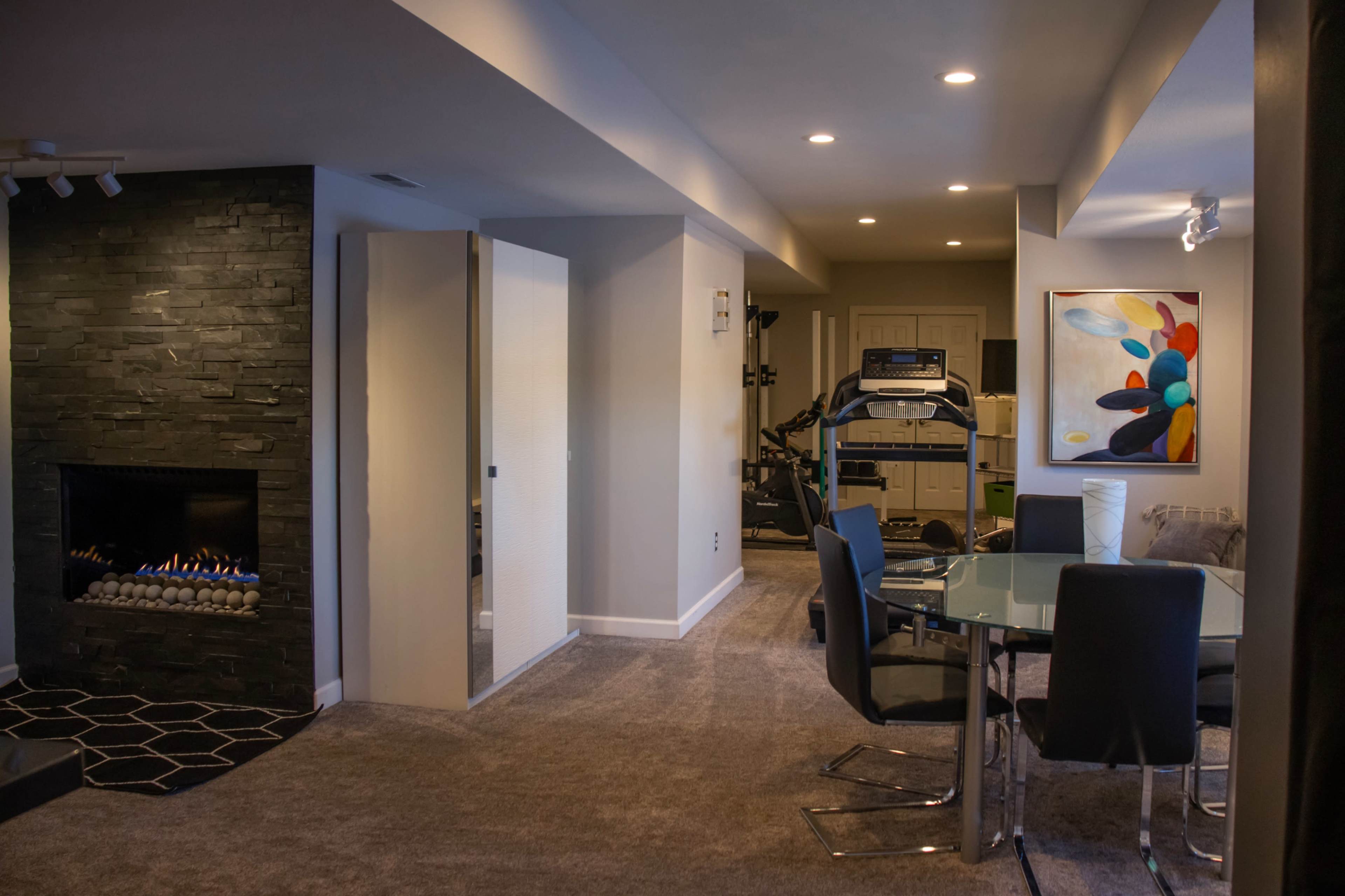 The image shows a modern living space featuring a glass dining table with dark chairs, a black stone fireplace, and an exercise area with equipment.