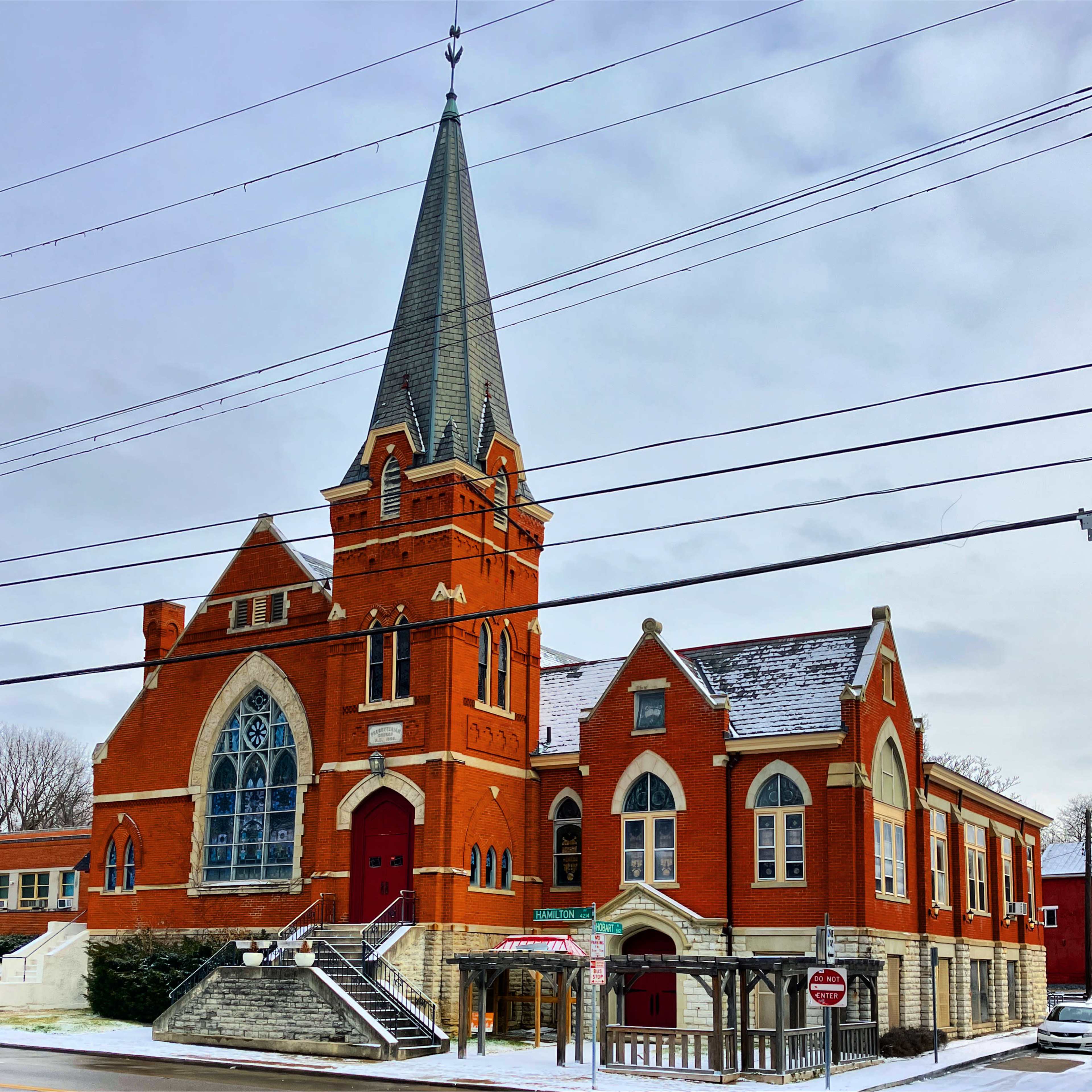 A brick church with a tall steeple and red doors stands at the corner of a snowy street, surrounded by power lines.