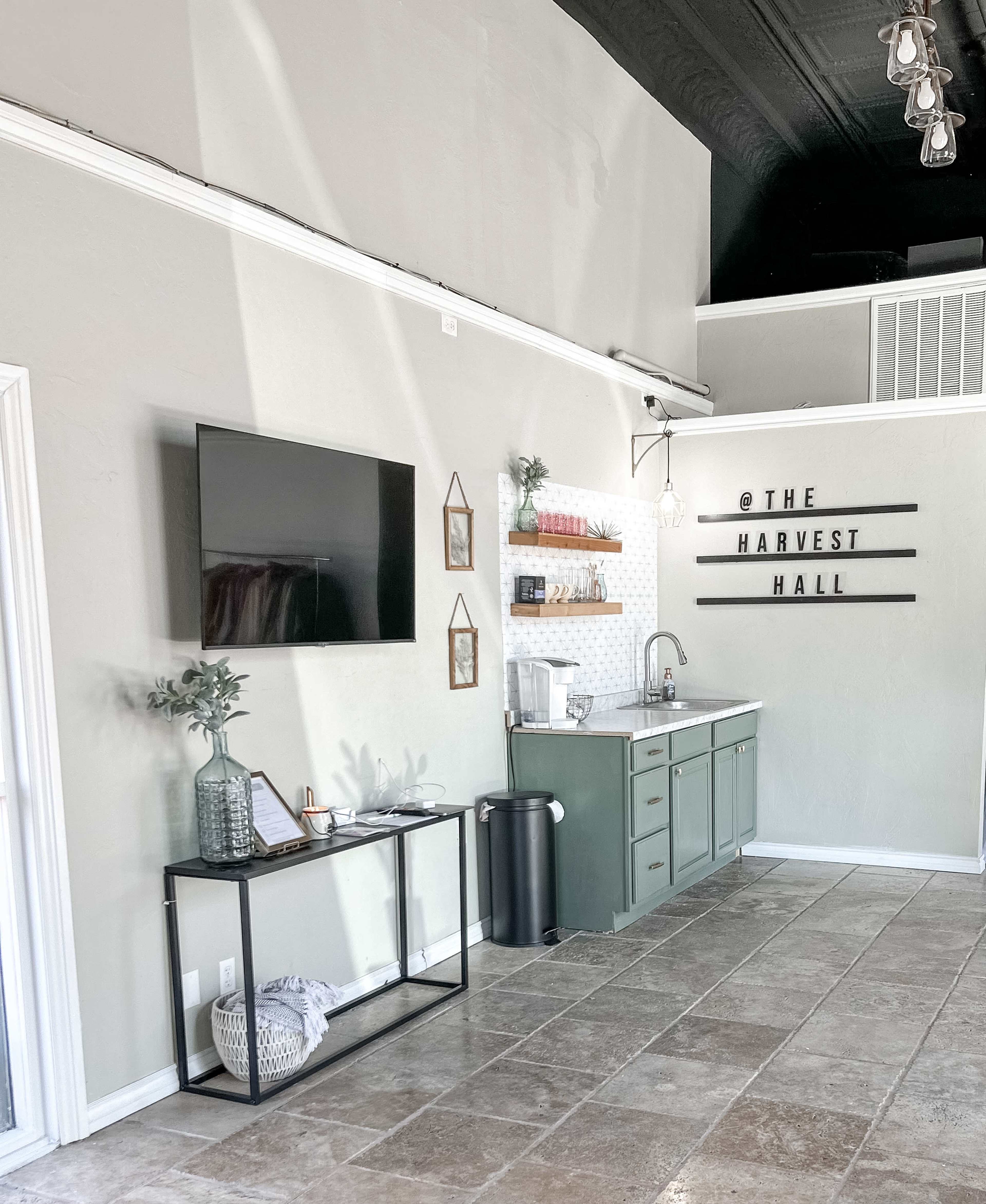 The image shows a modern interior space with a green kitchenette, a wall-mounted TV, and decorative elements including shelves and wall signage that reads "THE HARVEST HALL."