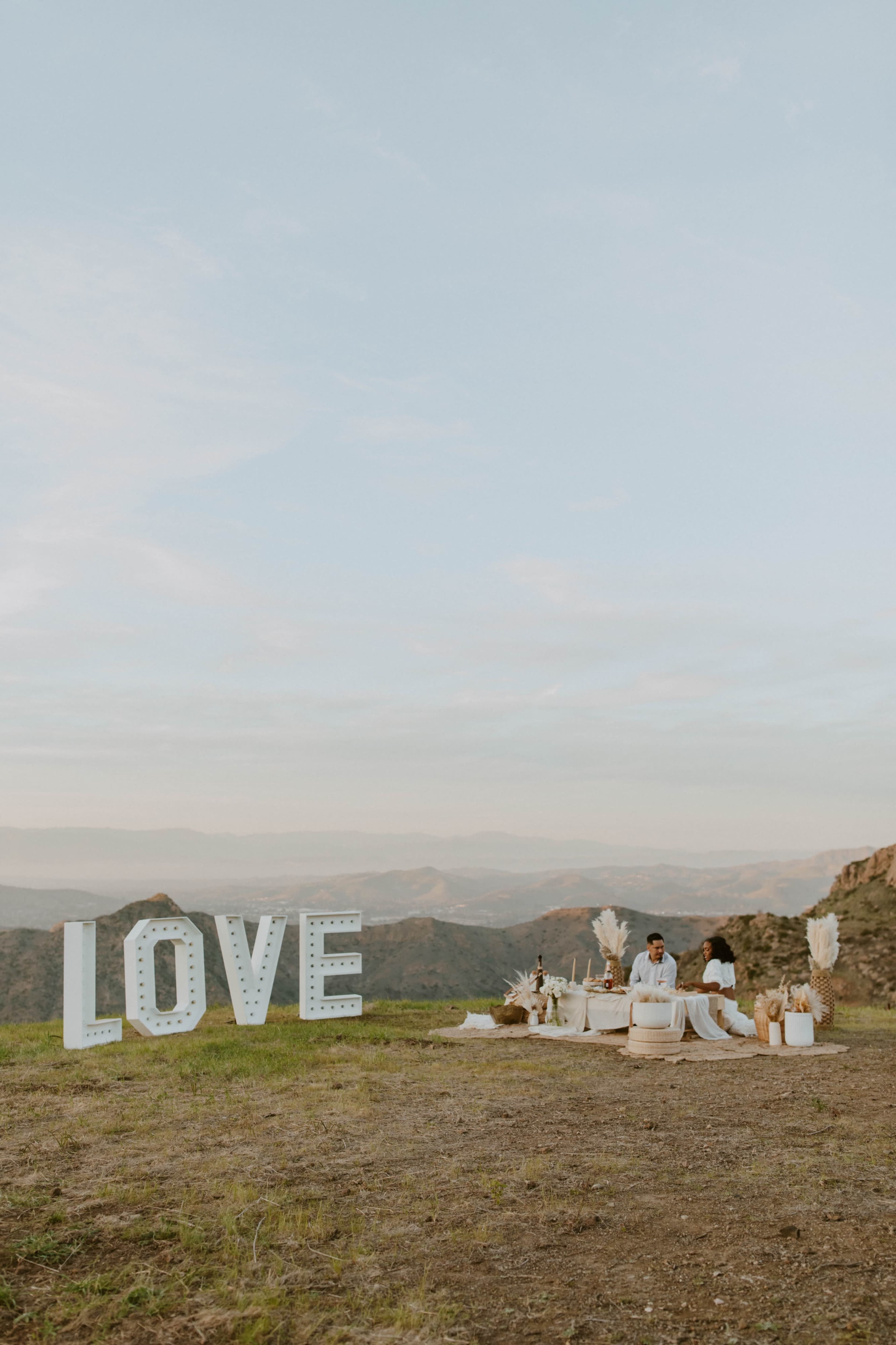A couple sits at a picnic setup in a scenic outdoor location, with large letters spelling "LOVE" nearby.