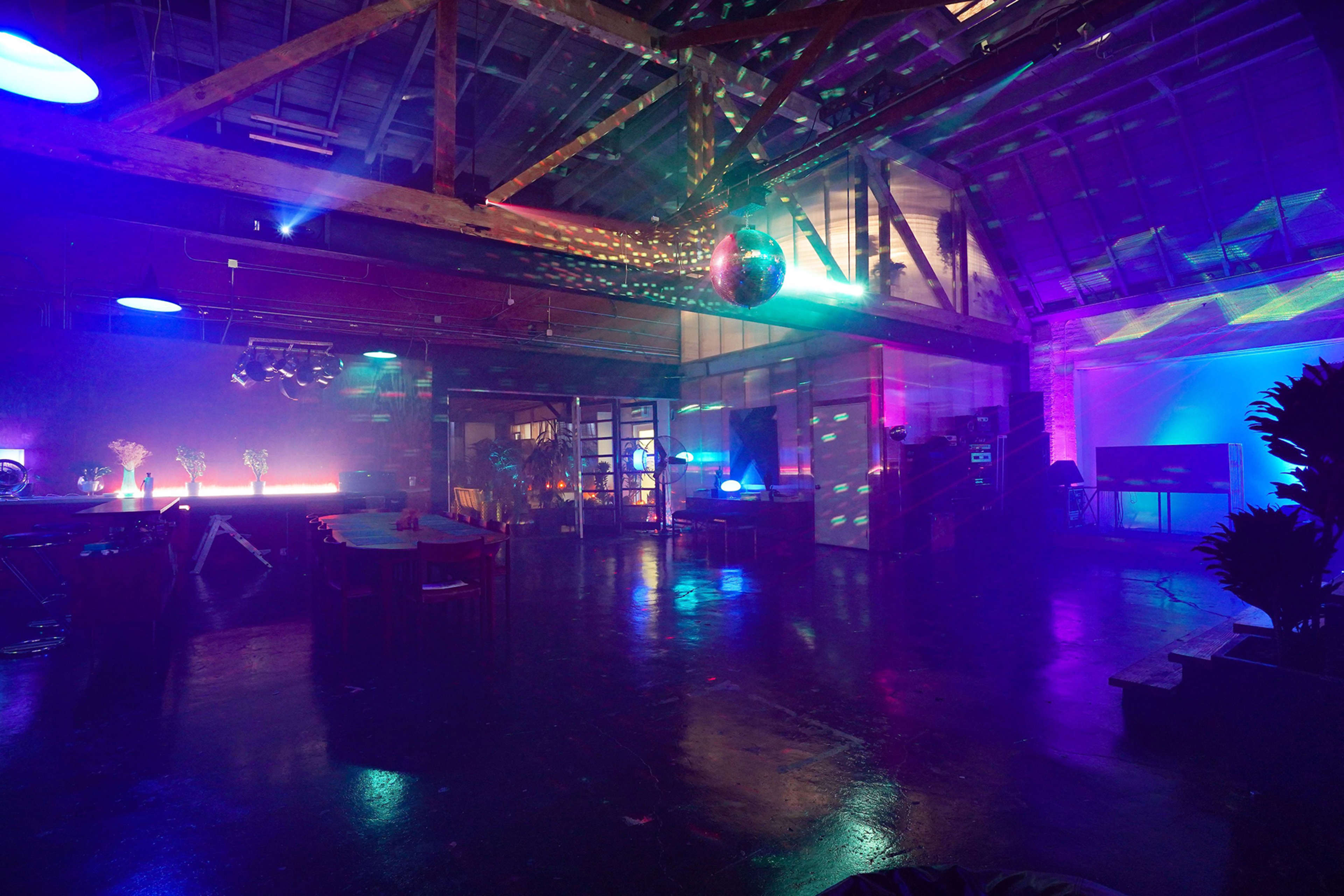 A spacious interior with colorful lighting effects, a disco ball, and various event setups in a warehouse-like venue.