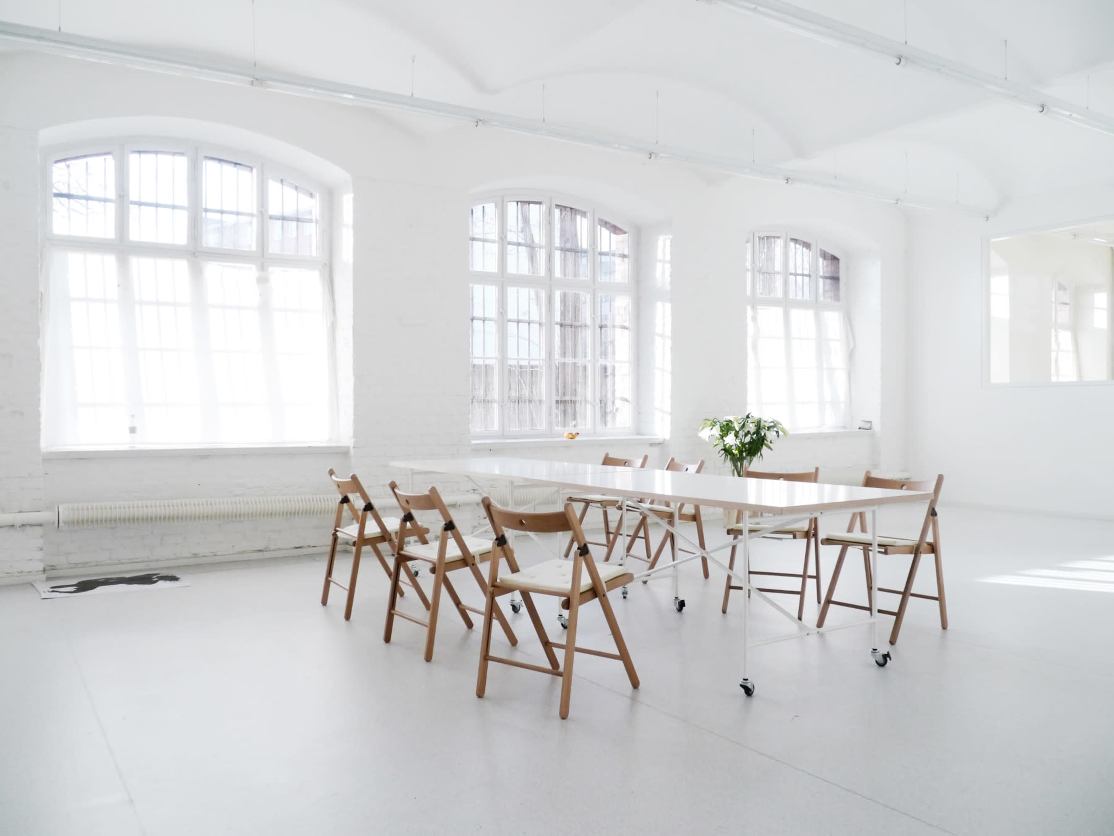 A spacious, bright room features a long white table surrounded by wooden folding chairs and large windows.