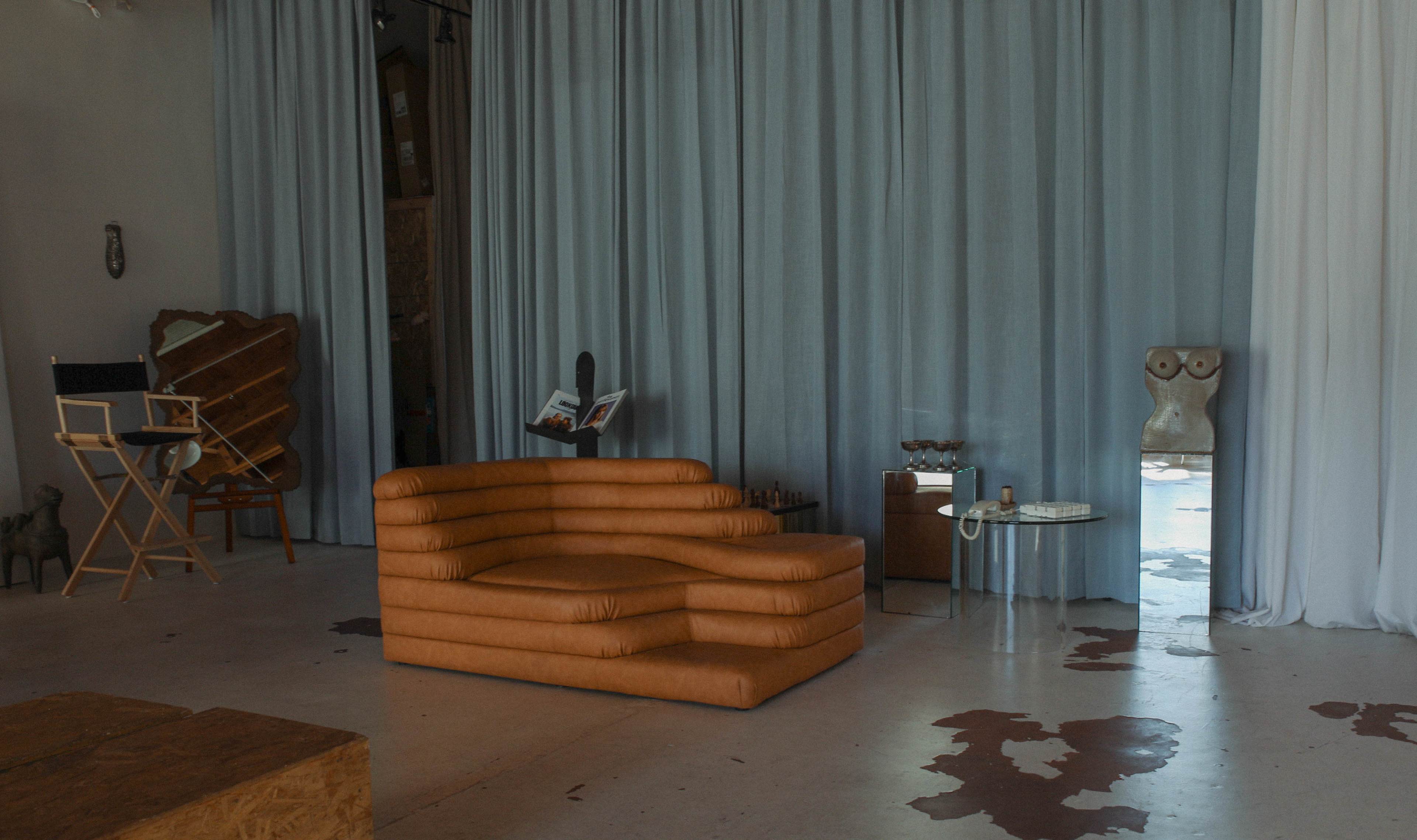 The image shows a modern interior featuring a curved brown couch, draped curtains, and various decorative objects in a minimalist space.
