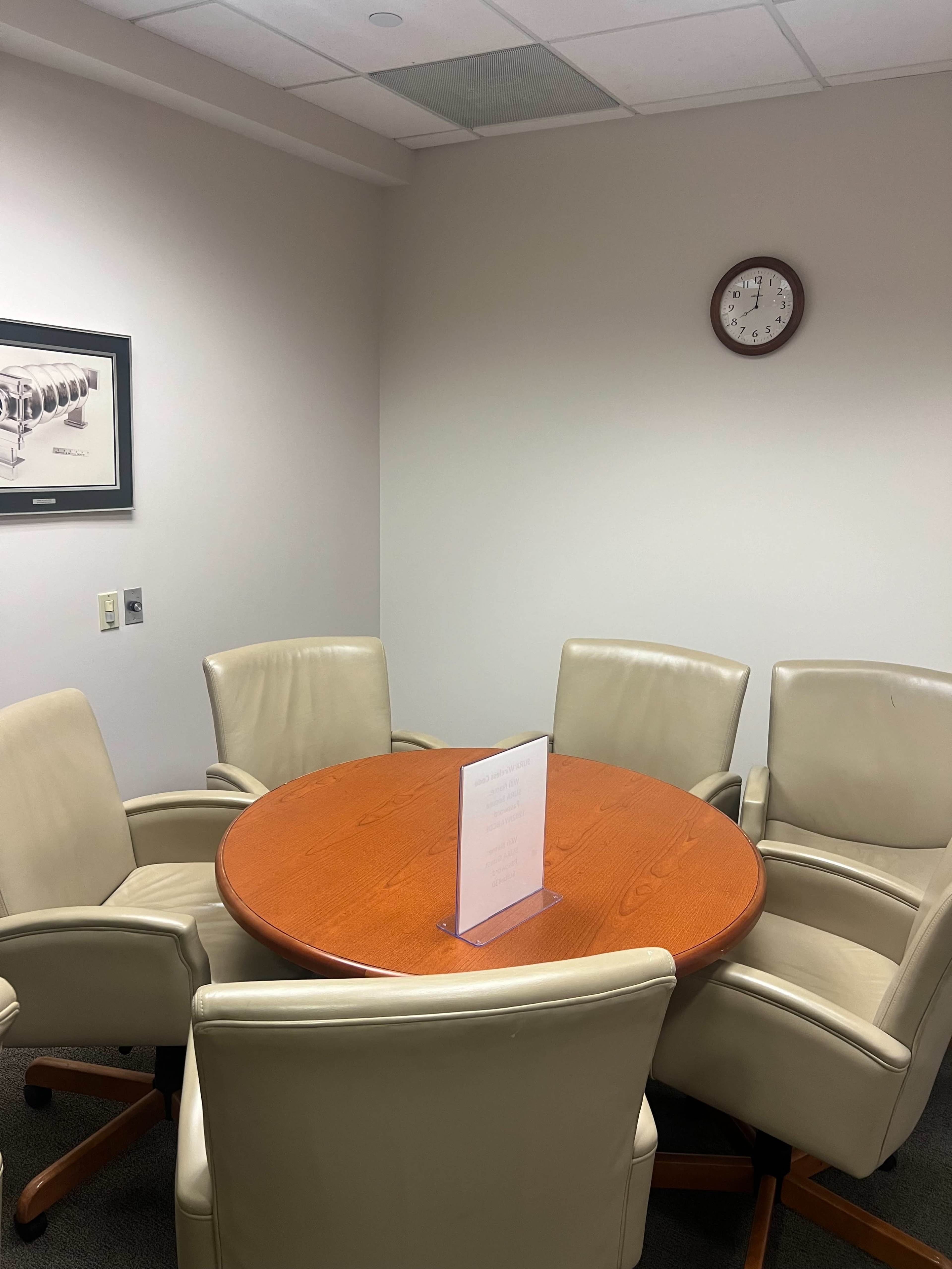 A round wooden table is surrounded by four light-colored chairs in a small meeting room, with a clock and a framed picture on the walls.