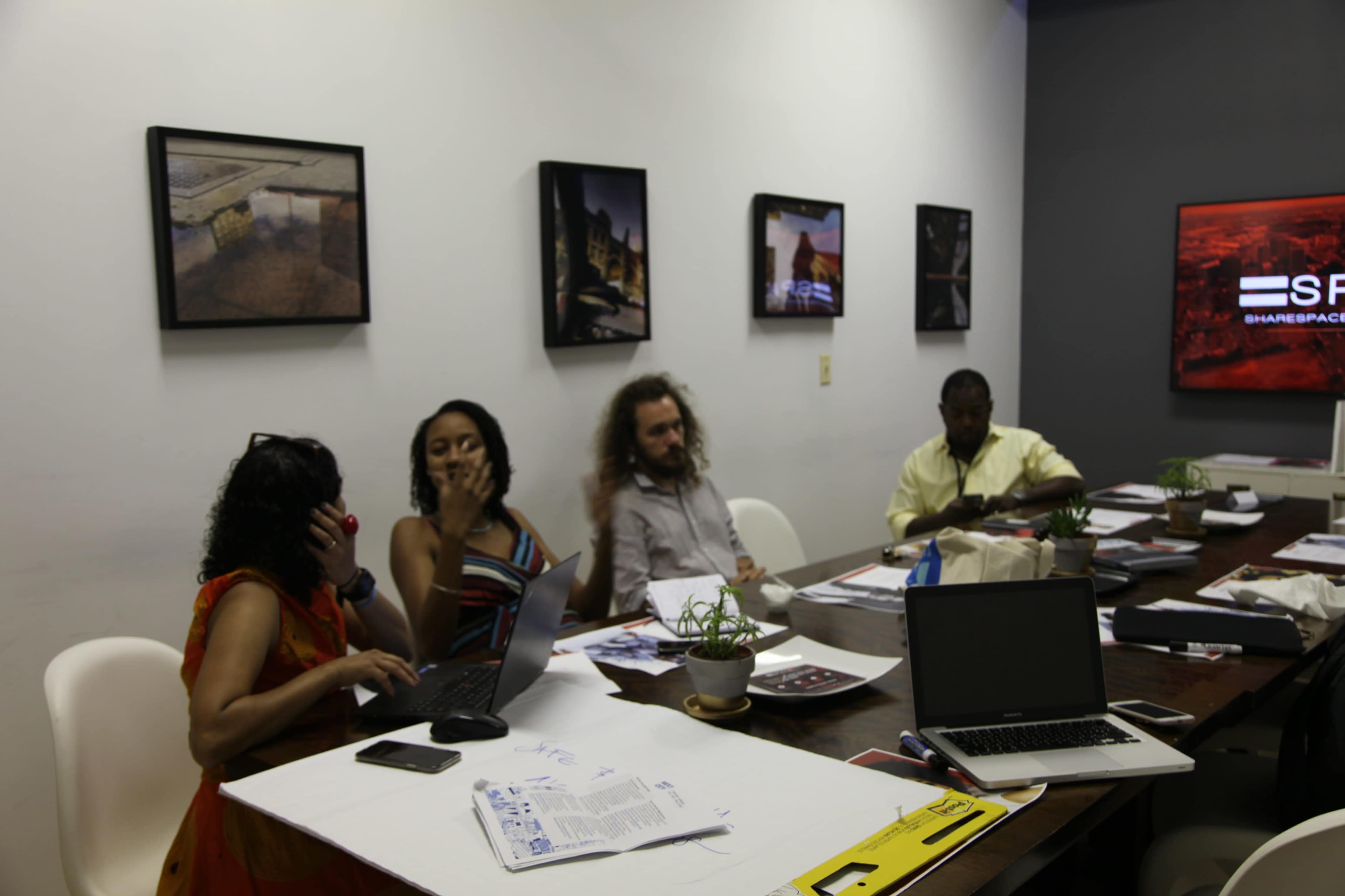 A group of four people is seated around a table in an office, working on laptops and discussing documents, while artwork hangs on the walls.