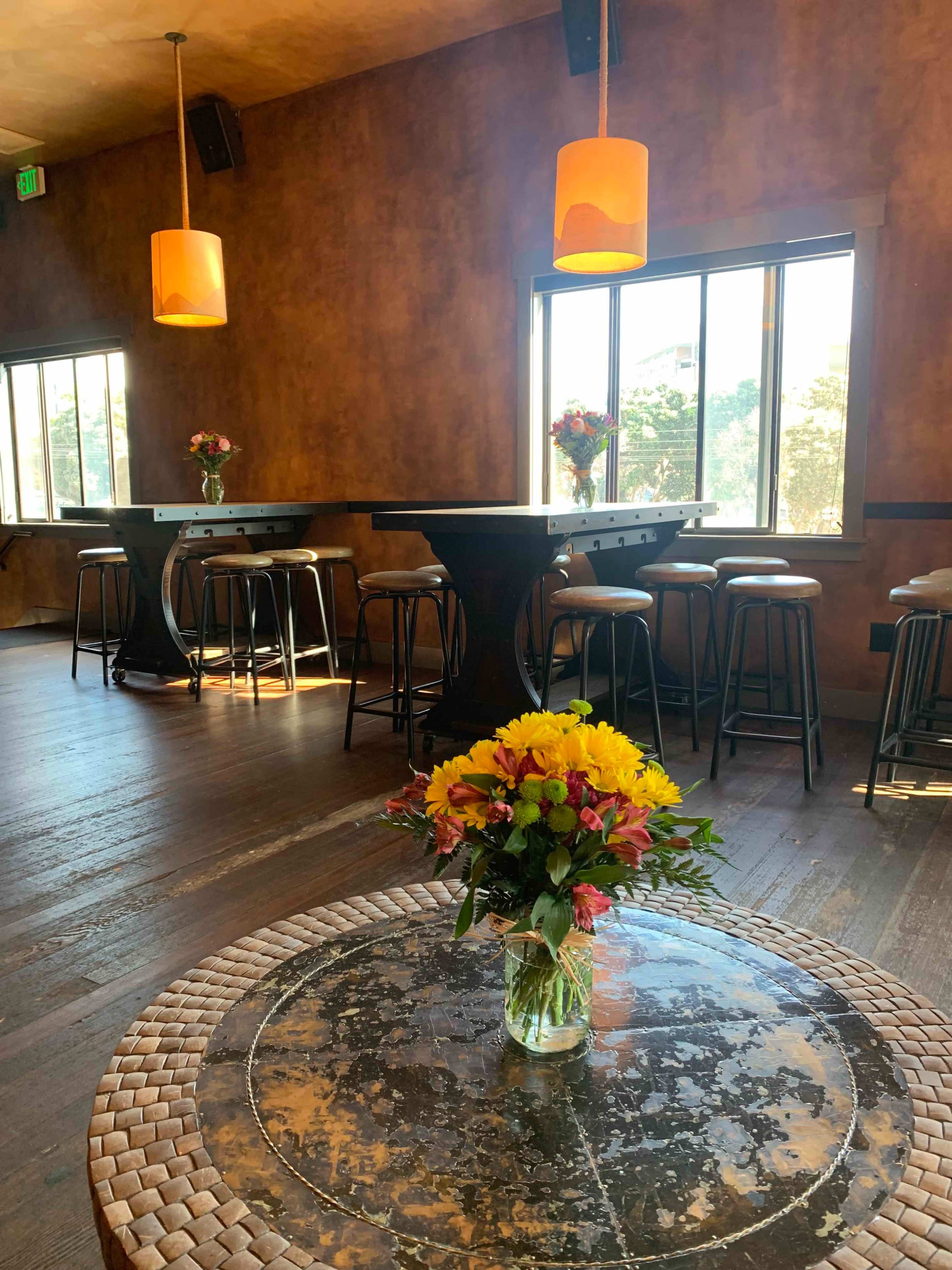 A spacious interior of a bar or restaurant featuring wooden floors, dark tables, high stools, and a floral arrangement on a circular table.