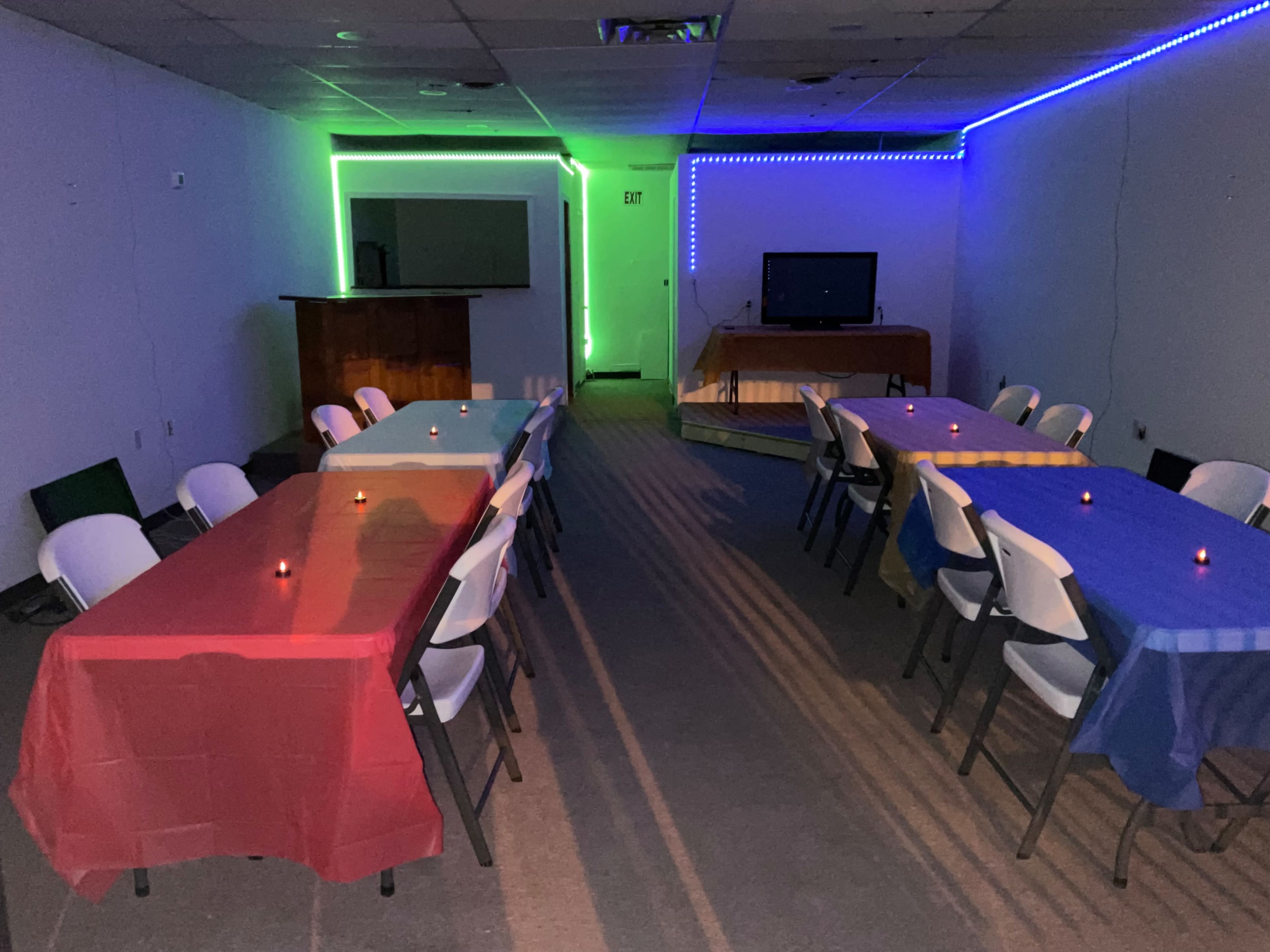 The image shows an empty room with several tables covered in red and blue tablecloths, surrounded by ambient lighting and a television in the background.