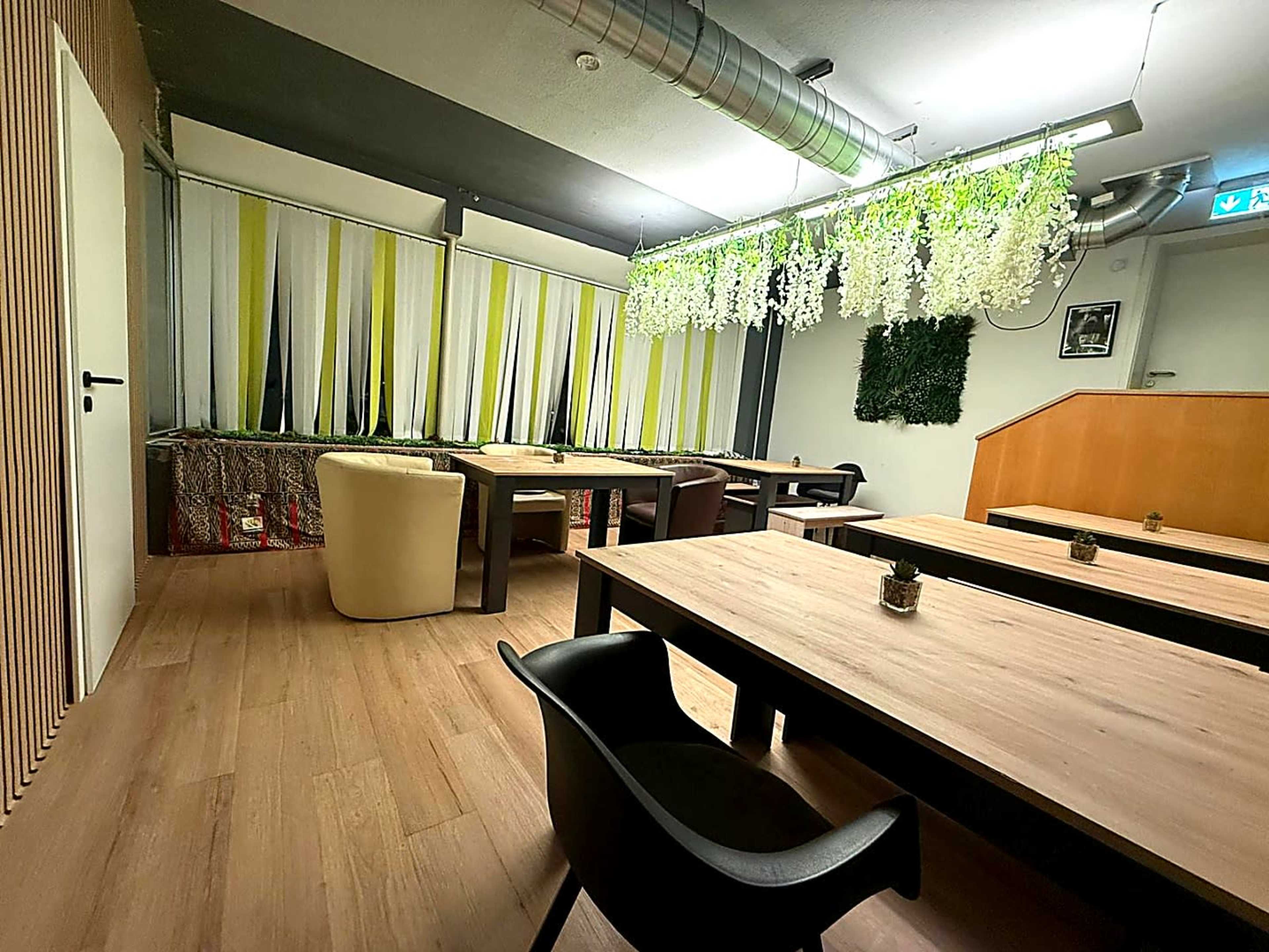 The image shows a modern, minimalist interior of a restaurant or café, featuring wooden tables, black chairs, and decorative greenery hanging from the ceiling.
