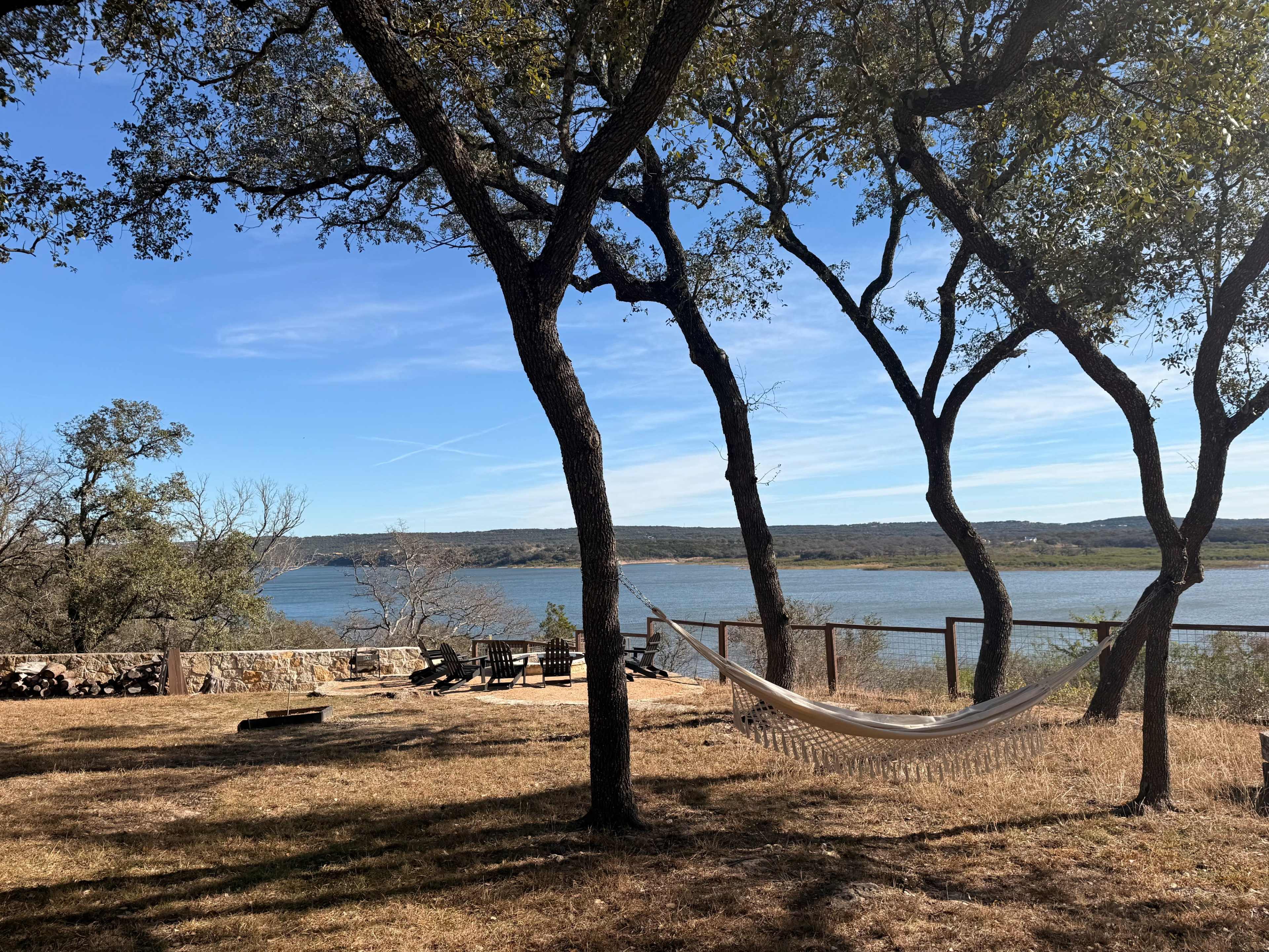 The image shows a serene lakeside scene with a hammock suspended between two trees near a water body, surrounded by dry grass and scattered stone seating.