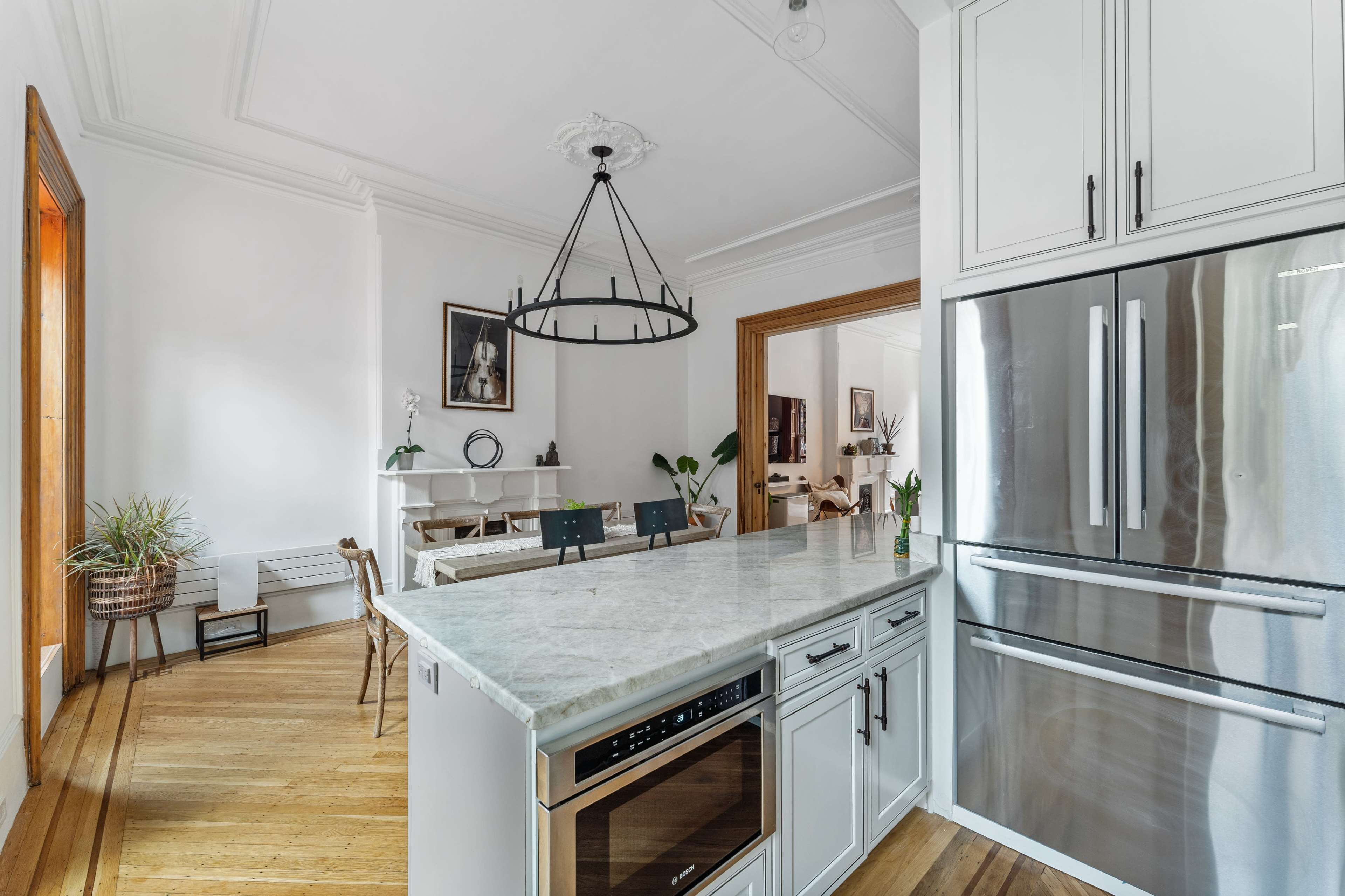 The image shows a modern kitchen with a marble island, stainless steel appliances, and a view into an adjacent dining area featuring wooden flooring and plants.