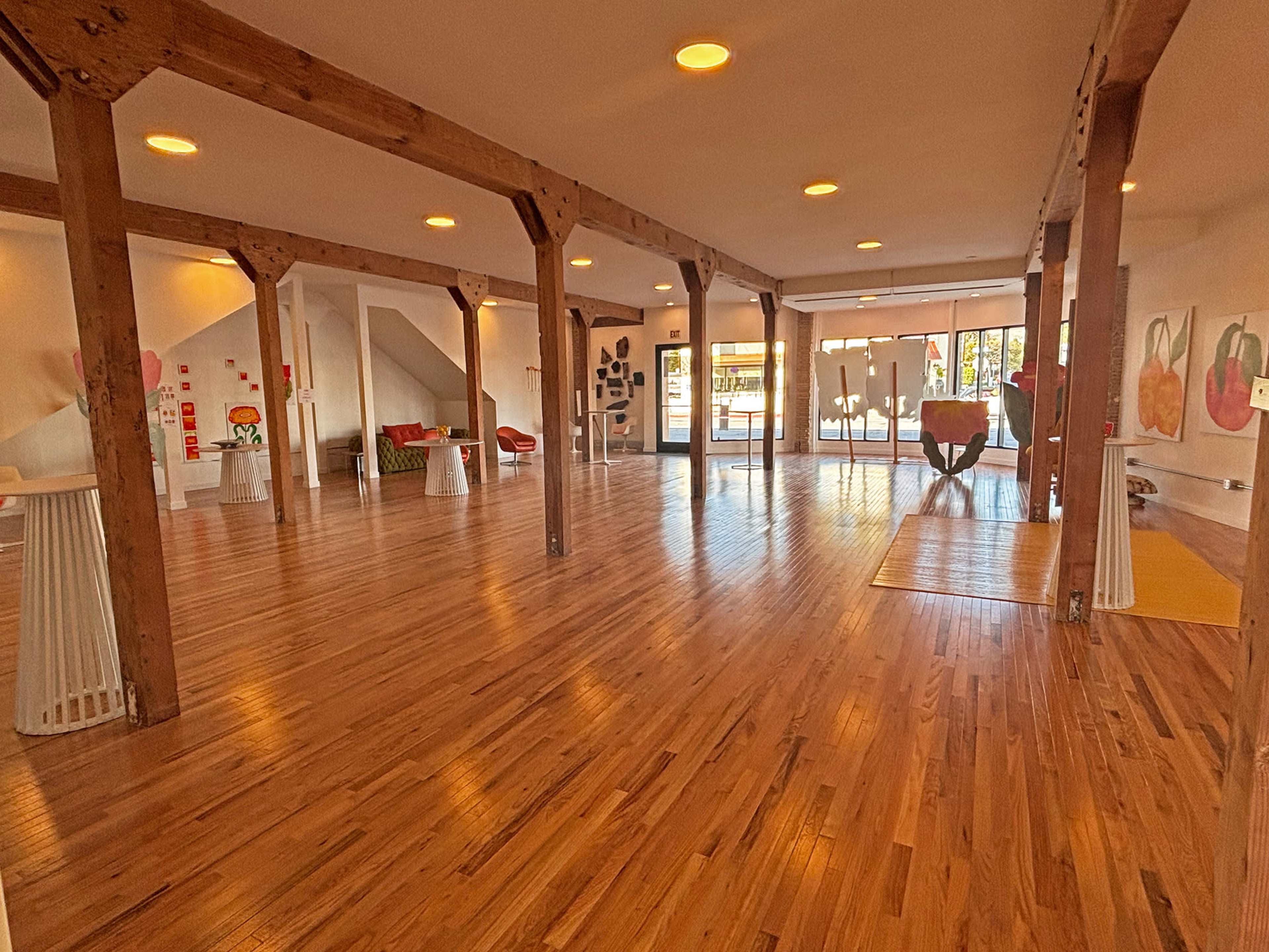 The image shows an empty, spacious room with wooden floors, exposed beams, and large windows allowing natural light to enter.