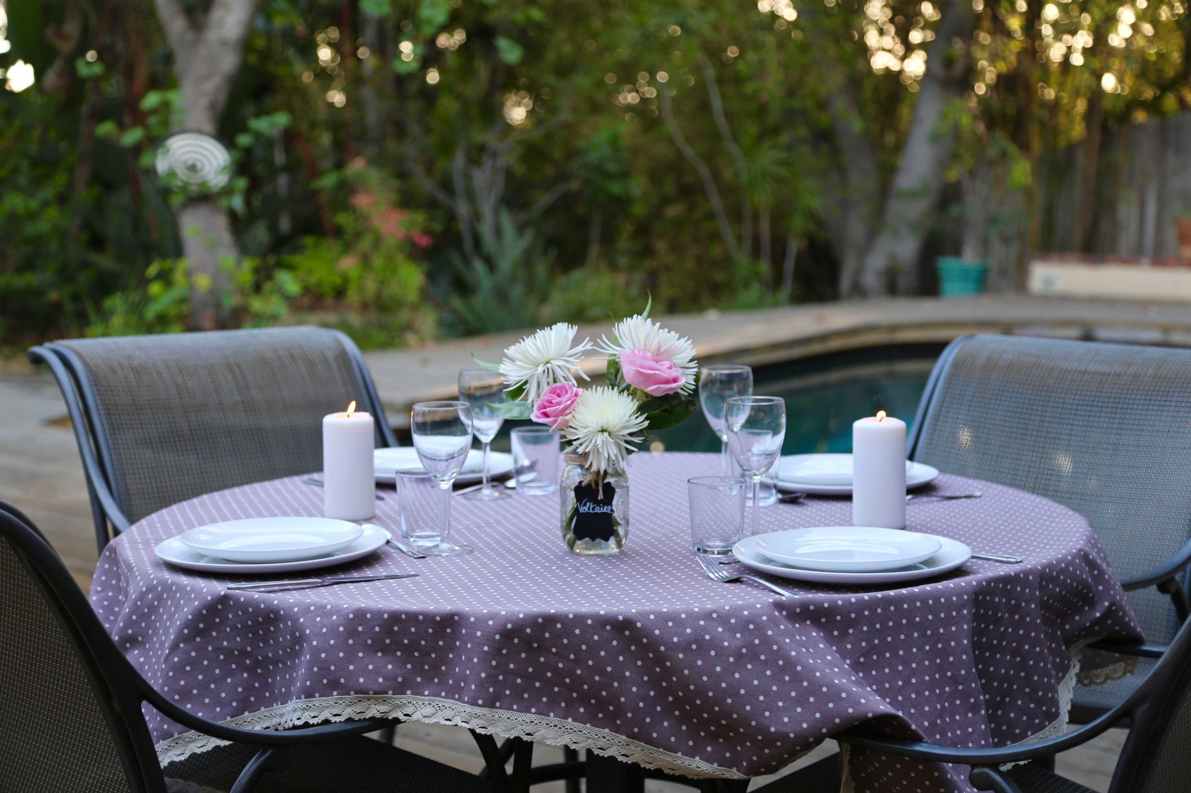 A round table with a polka dot tablecloth is set for dinner outdoors, featuring a floral centerpiece, candles, and diningware next to a pool.