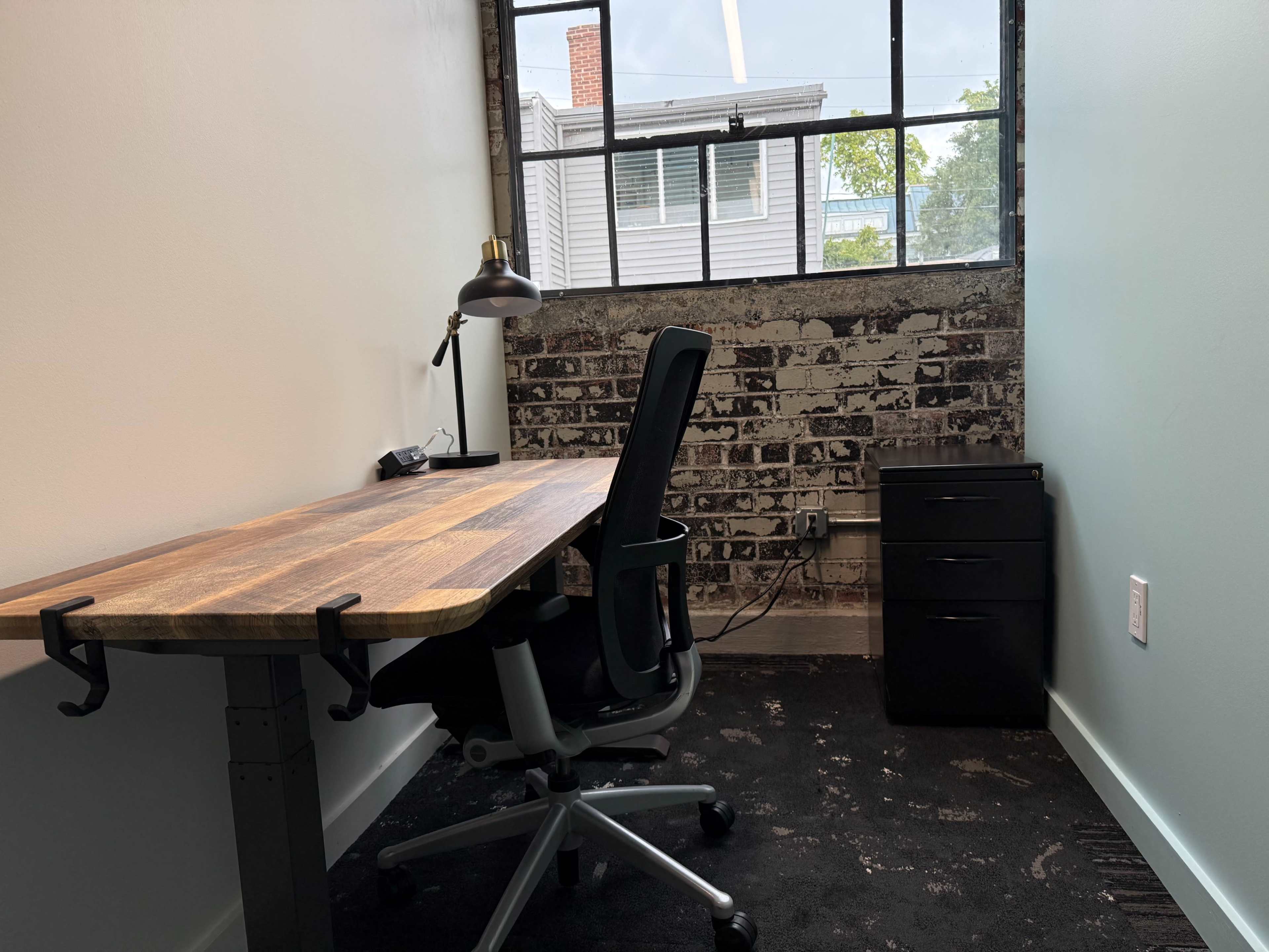 The image shows a minimalist office space featuring a wooden desk, an office chair, a black filing cabinet, and a large window with a view of a building and trees outside.