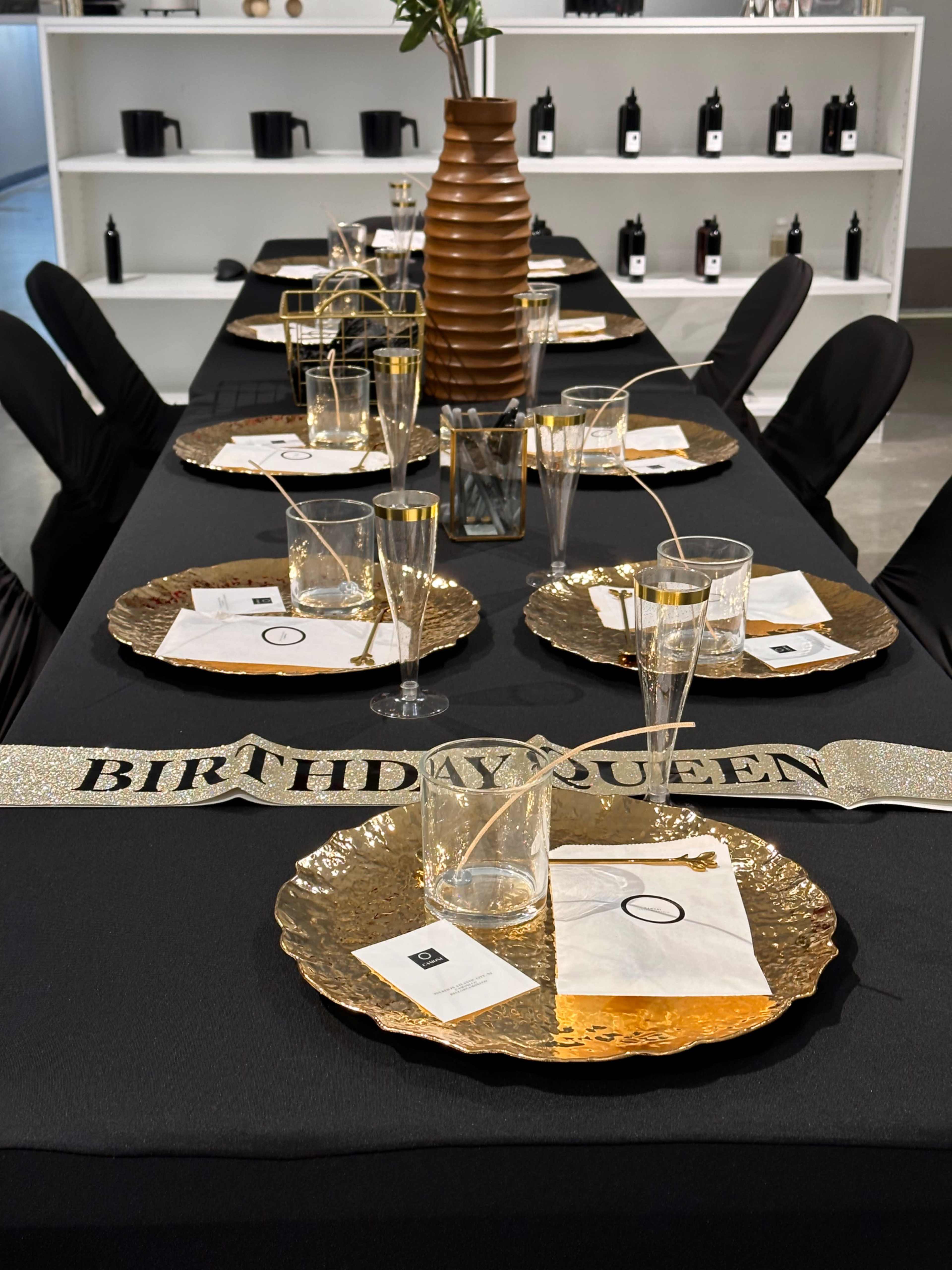 A black table is set with gold-rimmed plates, clear glasses, and a "BIRTHDAY QUEEN" banner, adorned with a decorative vase and surrounded by black chairs.