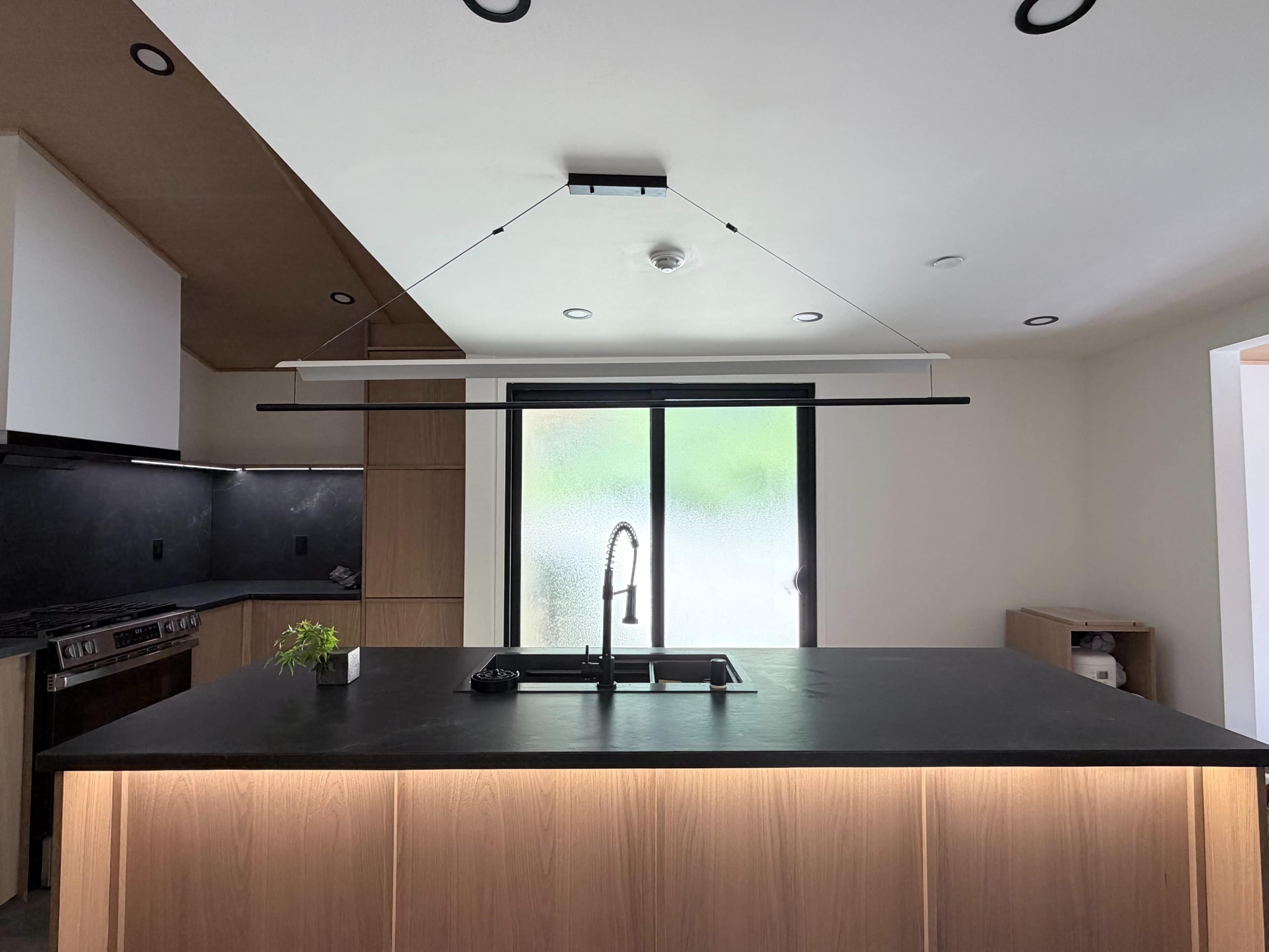 A modern kitchen features a central island with a black countertop, a minimalist light fixture overhead, and a sliding glass door leading to an outdoor area.