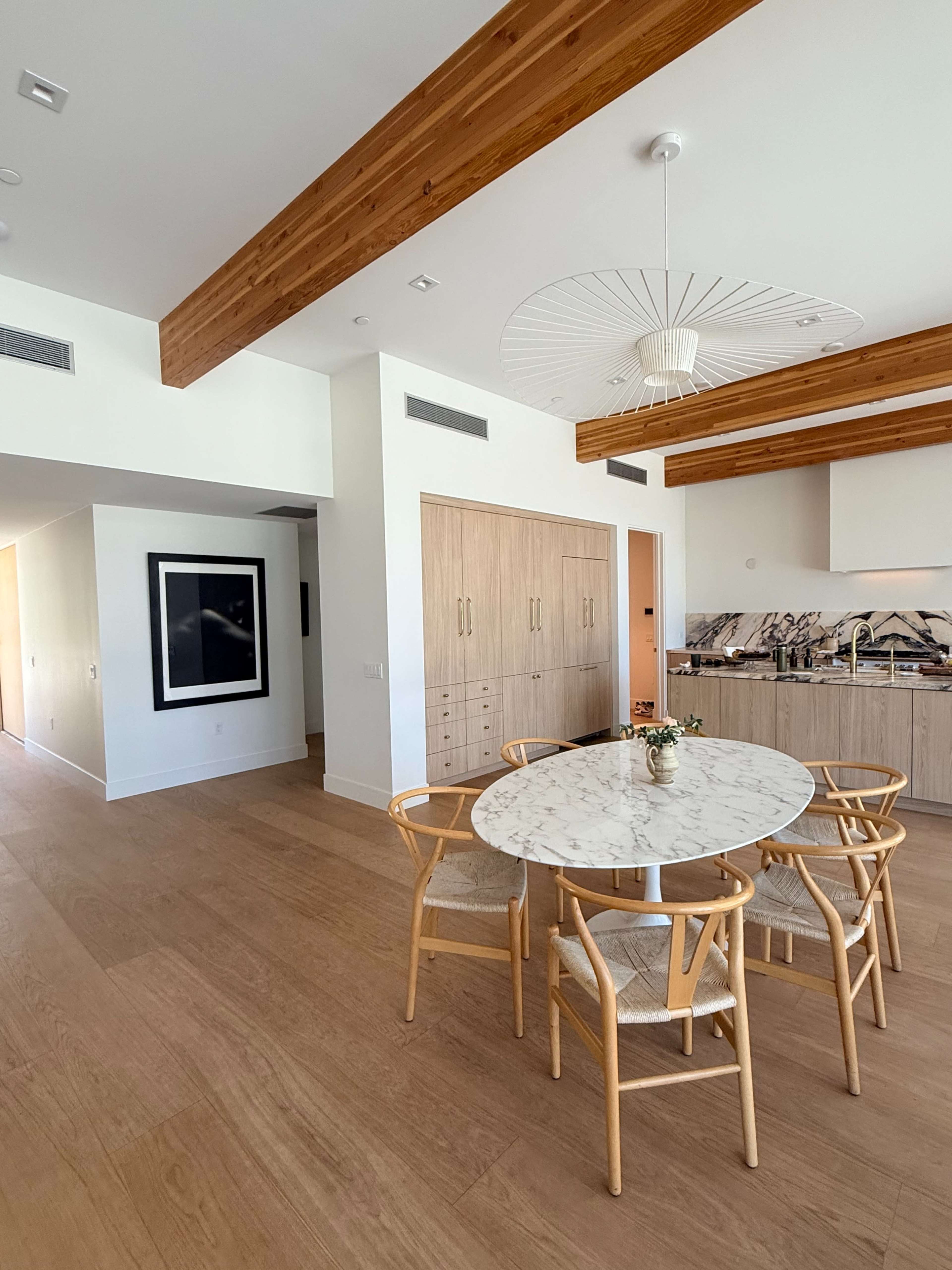 The image shows a modern kitchen and dining area with a circular marble table surrounded by wooden chairs and a backdrop of light wood beams and cabinetry.