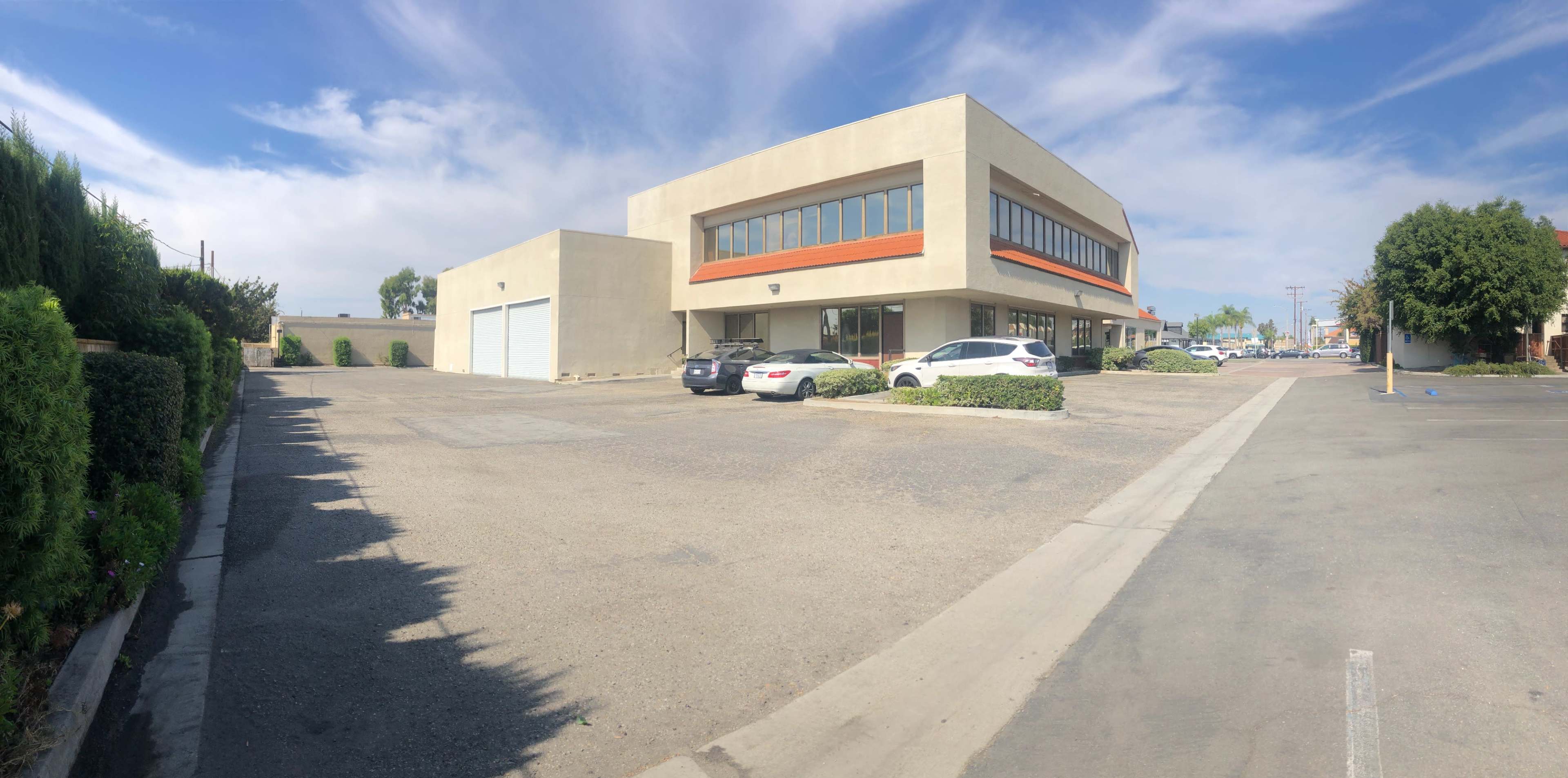 The image shows a two-story commercial building with large windows, surrounded by a gravel parking lot and several parked vehicles.