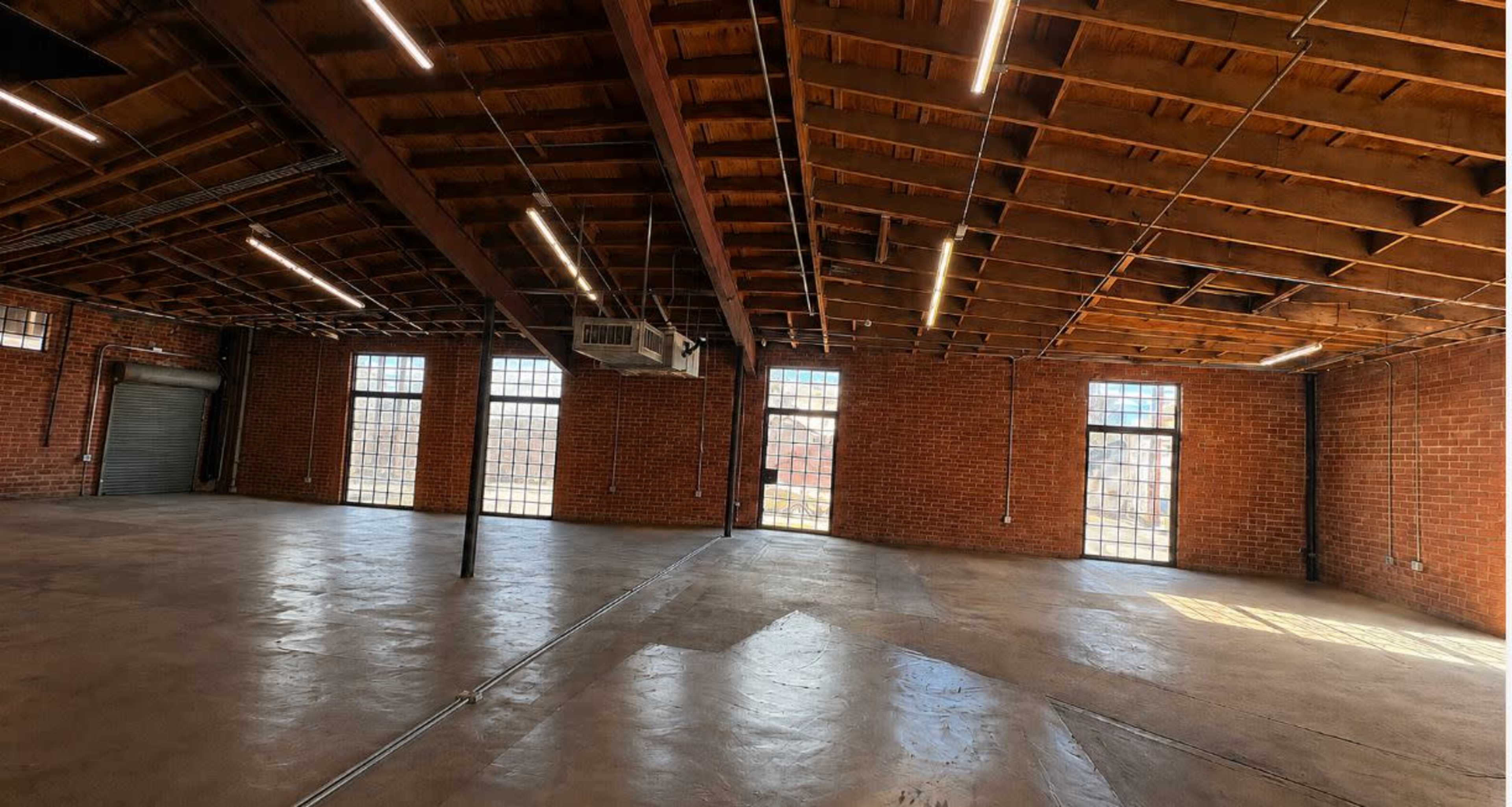 The image shows a large, empty industrial space with exposed brick walls, wooden beams, and large windows letting in natural light.