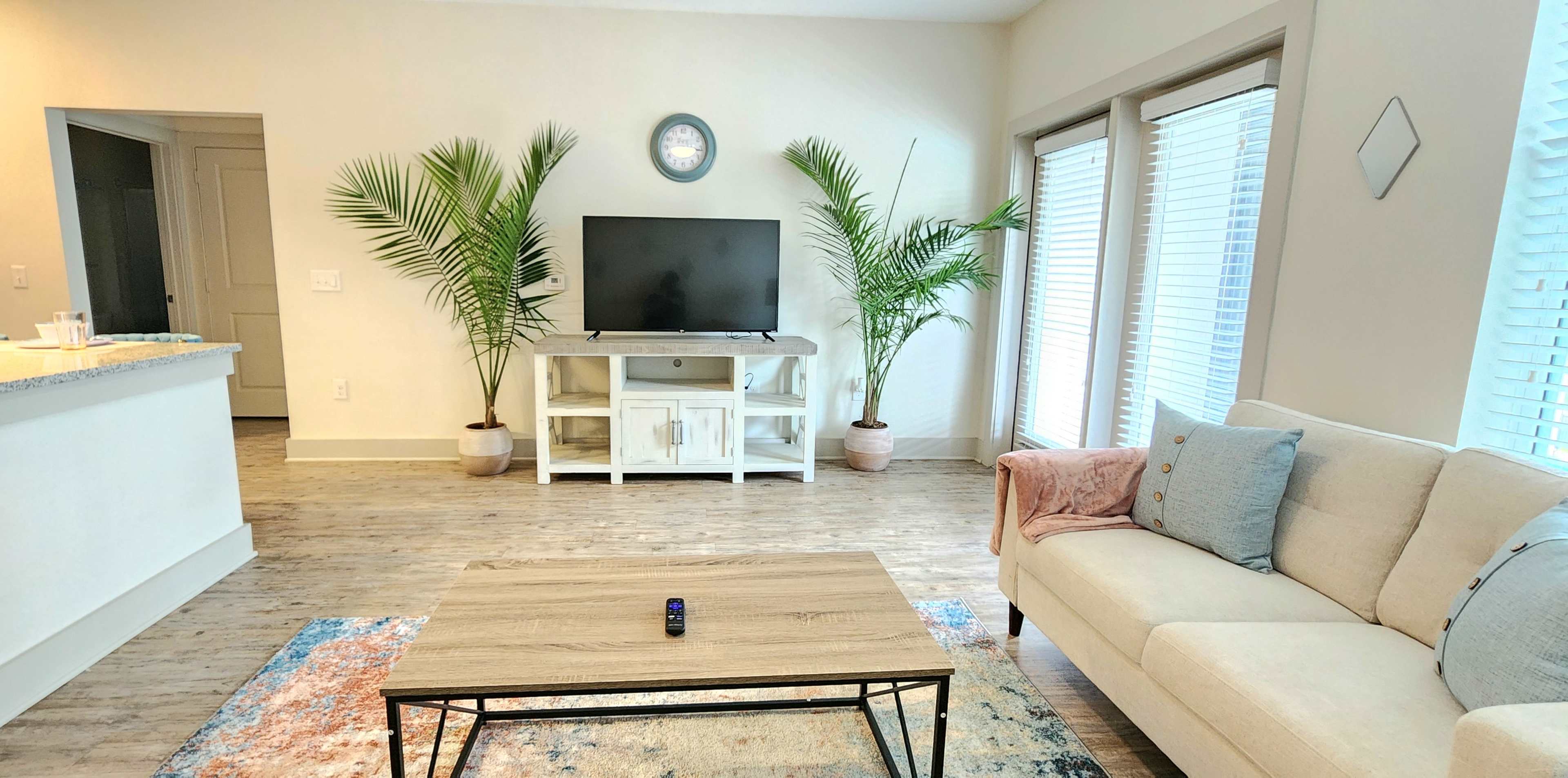 The living room features a light-colored couch, a wooden coffee table, a television on a white stand, and tropical plants near large windows.