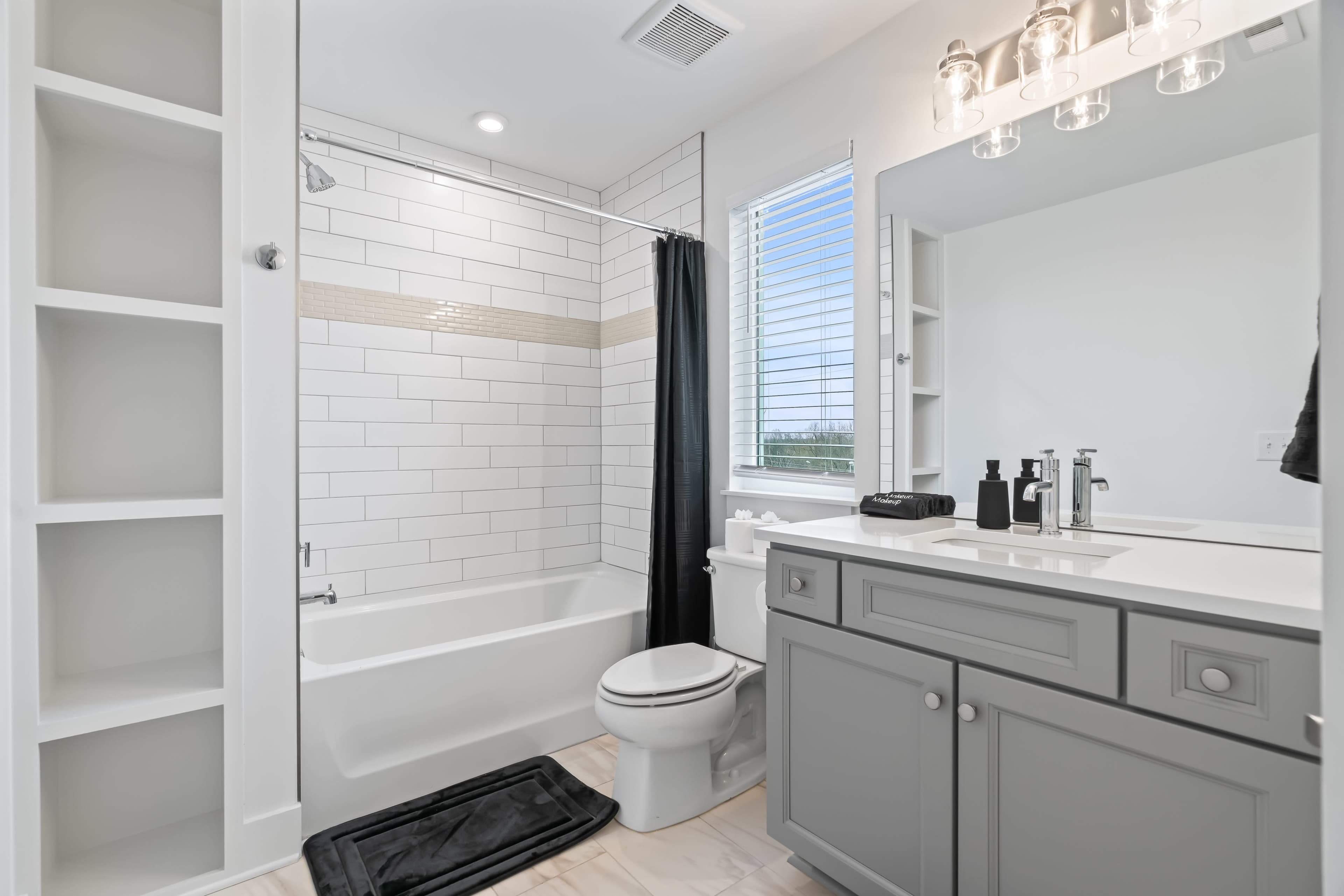 The bathroom features a white tiled shower with a black curtain, a gray vanity with a sink, and built-in shelves beside a window.