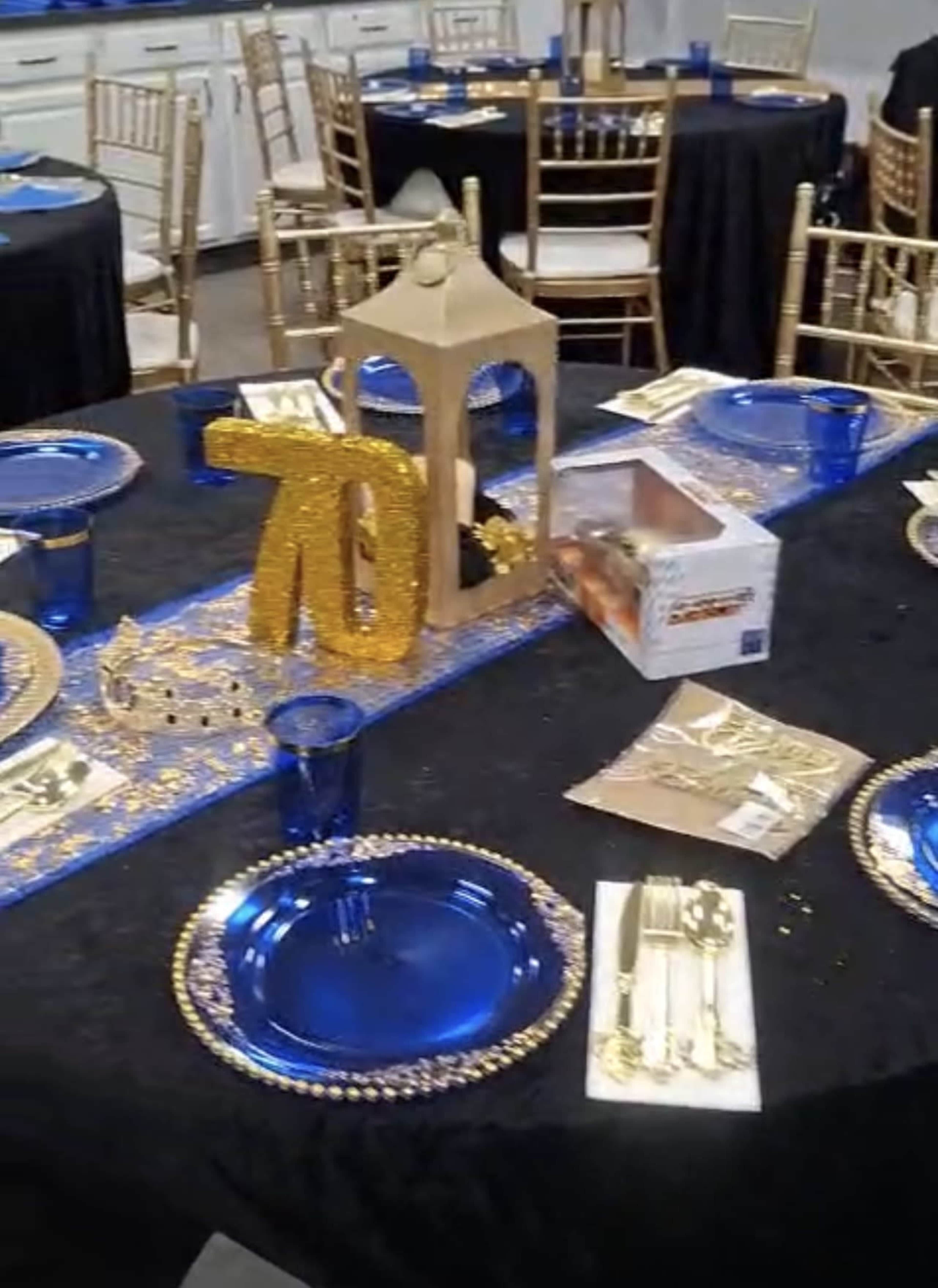 A banquet table is set with blue plates, golden cutlery, and a decorative centerpiece featuring a sparkling "70" on a black tablecloth.