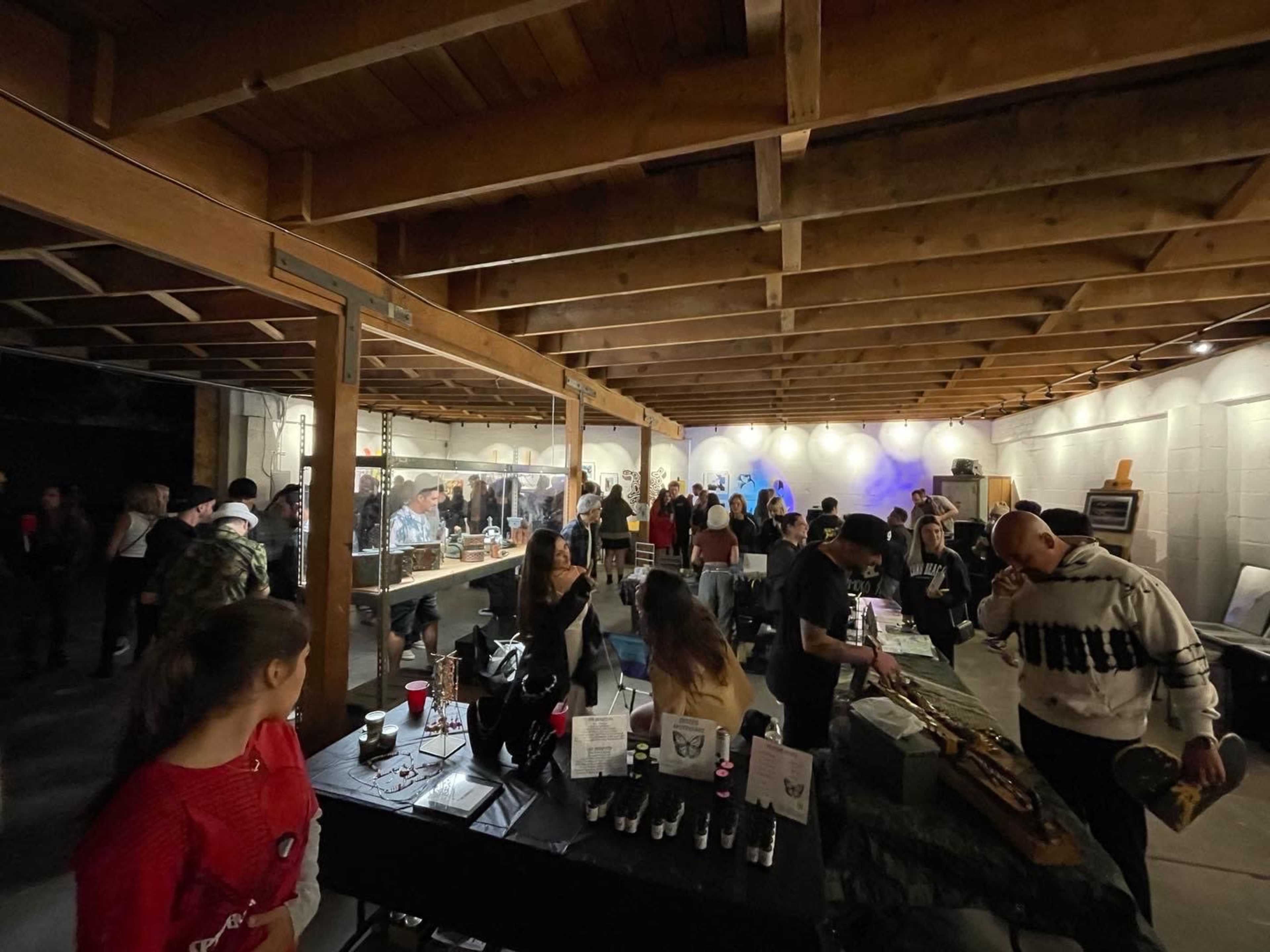 A crowd of people gathers in a spacious indoor area featuring tables displaying various items for sale amidst a backdrop of ambient lighting.