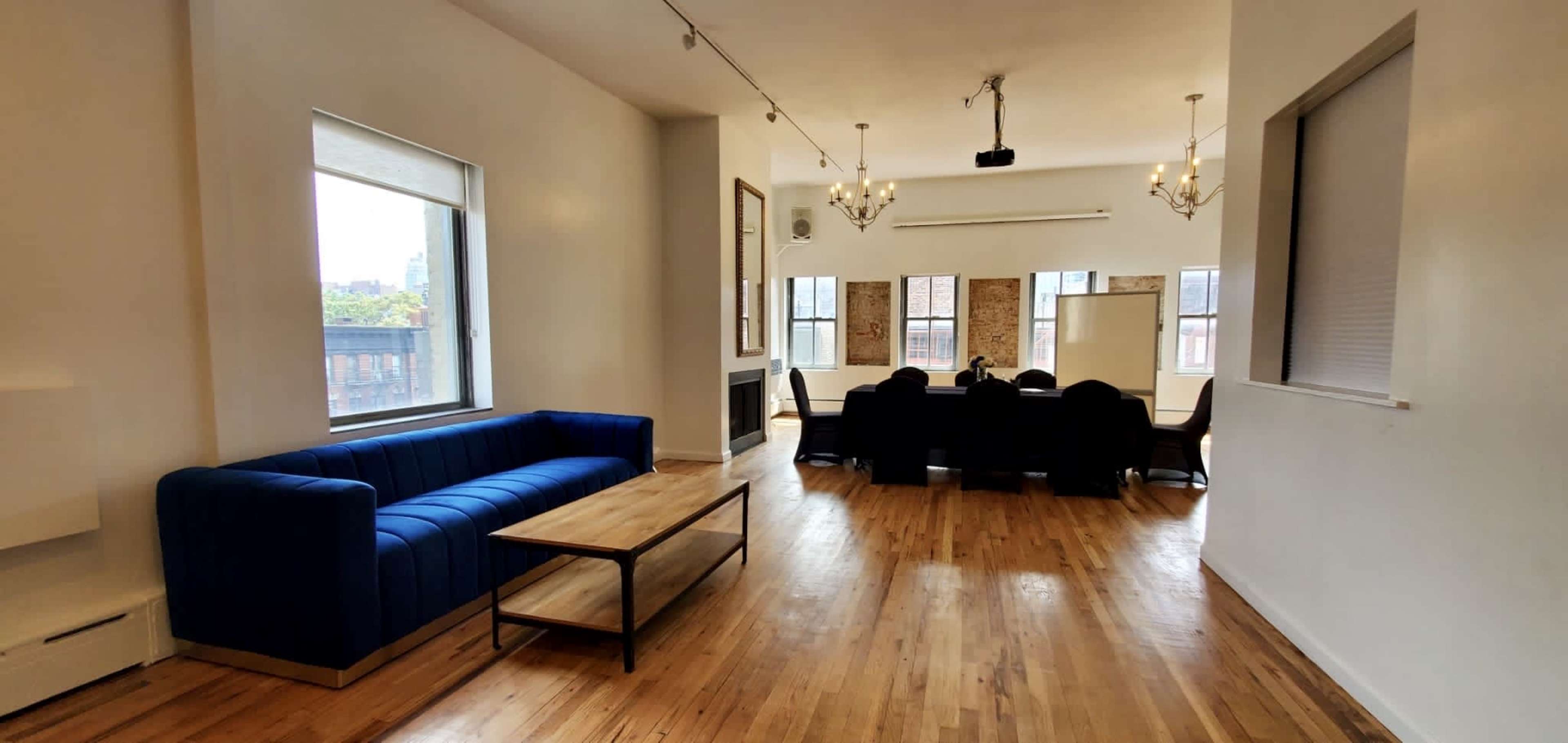 The image shows a spacious room with a blue couch and a wooden coffee table, featuring a conference area with a large table and chairs, illuminated by natural light through the windows.