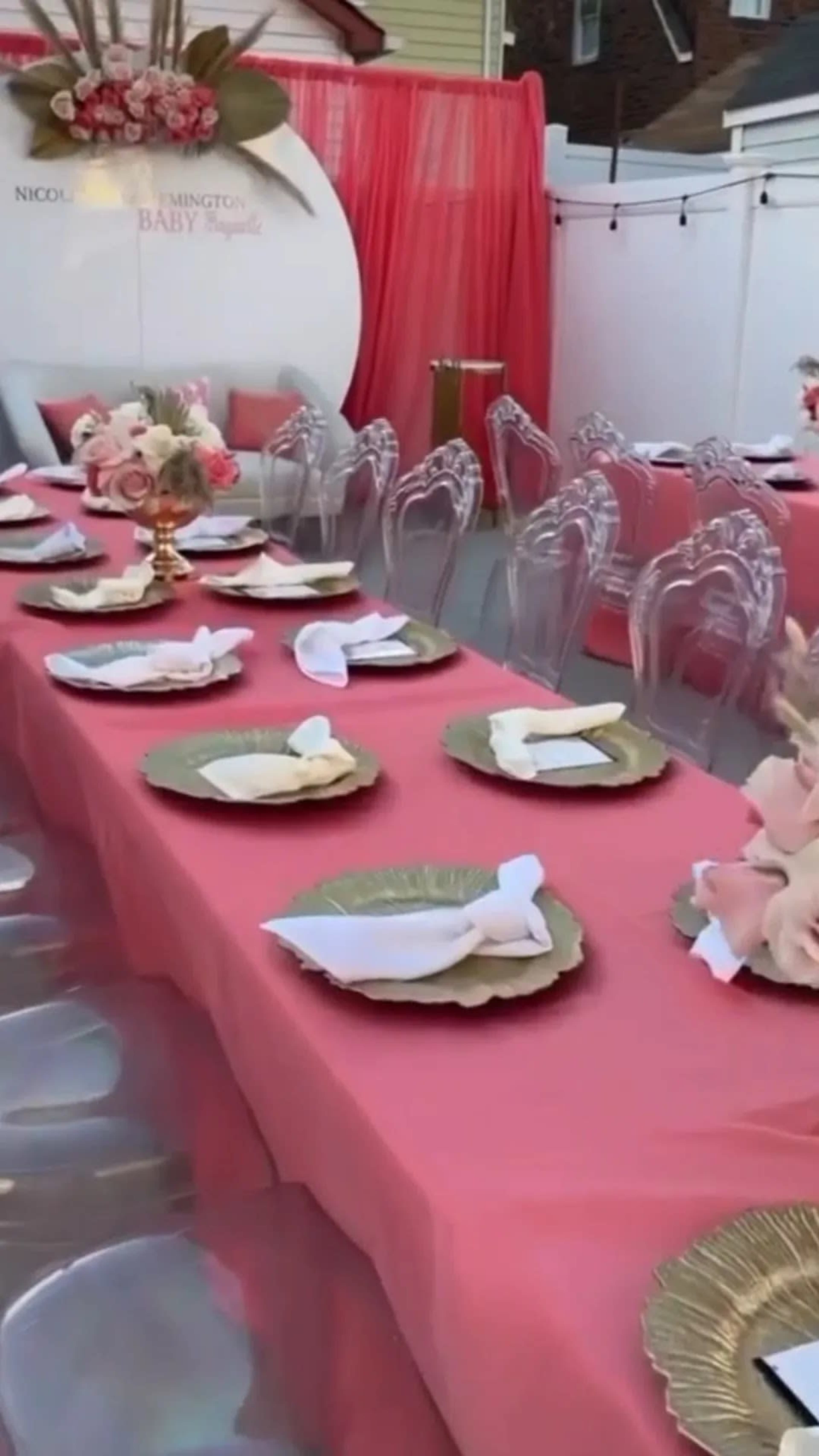 A long table is set with gold-rimmed plates and neatly folded napkins in an outdoor setting decorated with pink drapery and floral accents.