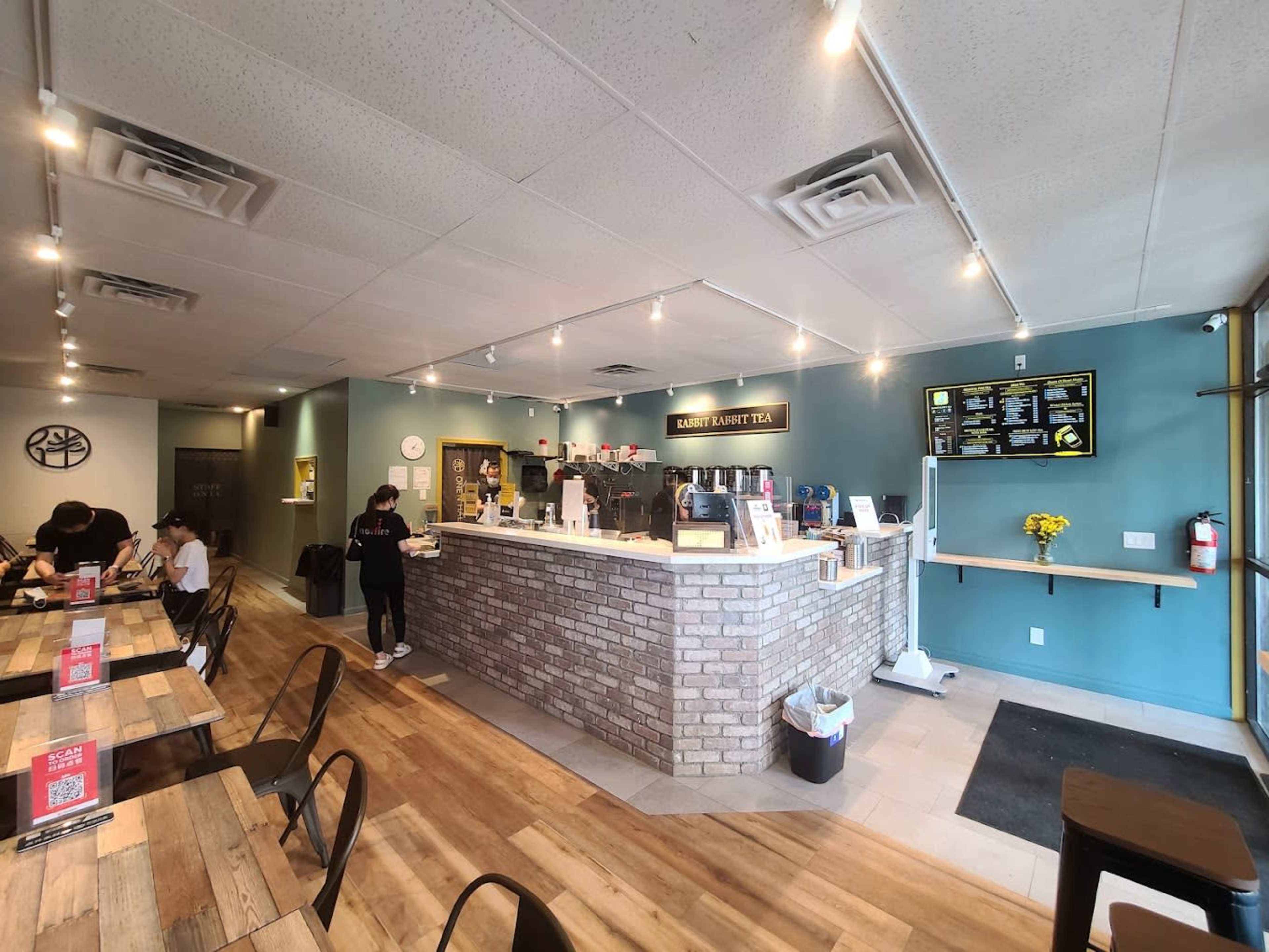 The interior of a tea shop featuring a service counter with a brick facade, tables arranged for customers, and a menu displayed on a wall.