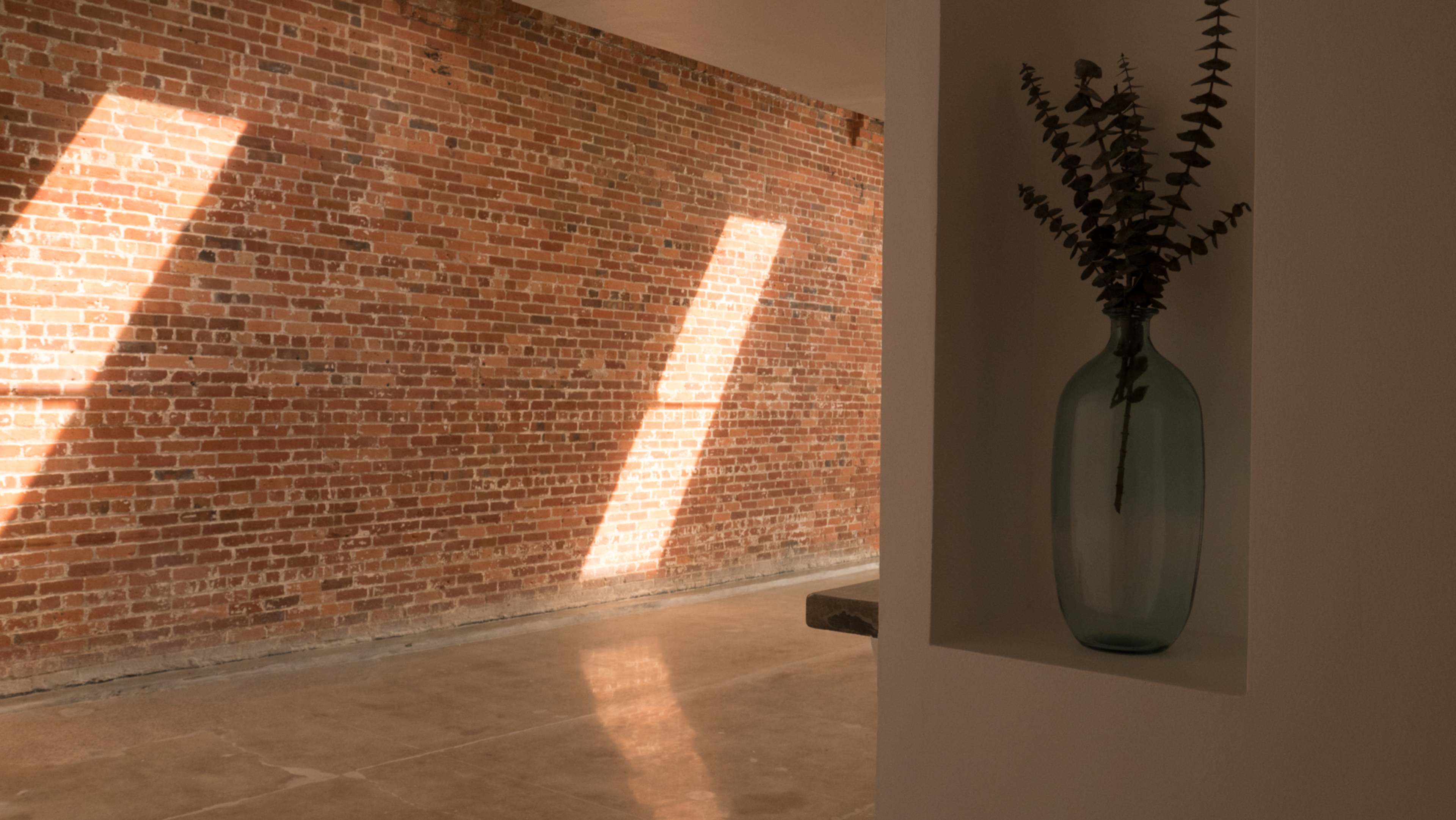 A clear vase holds eucalyptus branches on a shelf beside a sunlit brick wall and polished concrete floor.