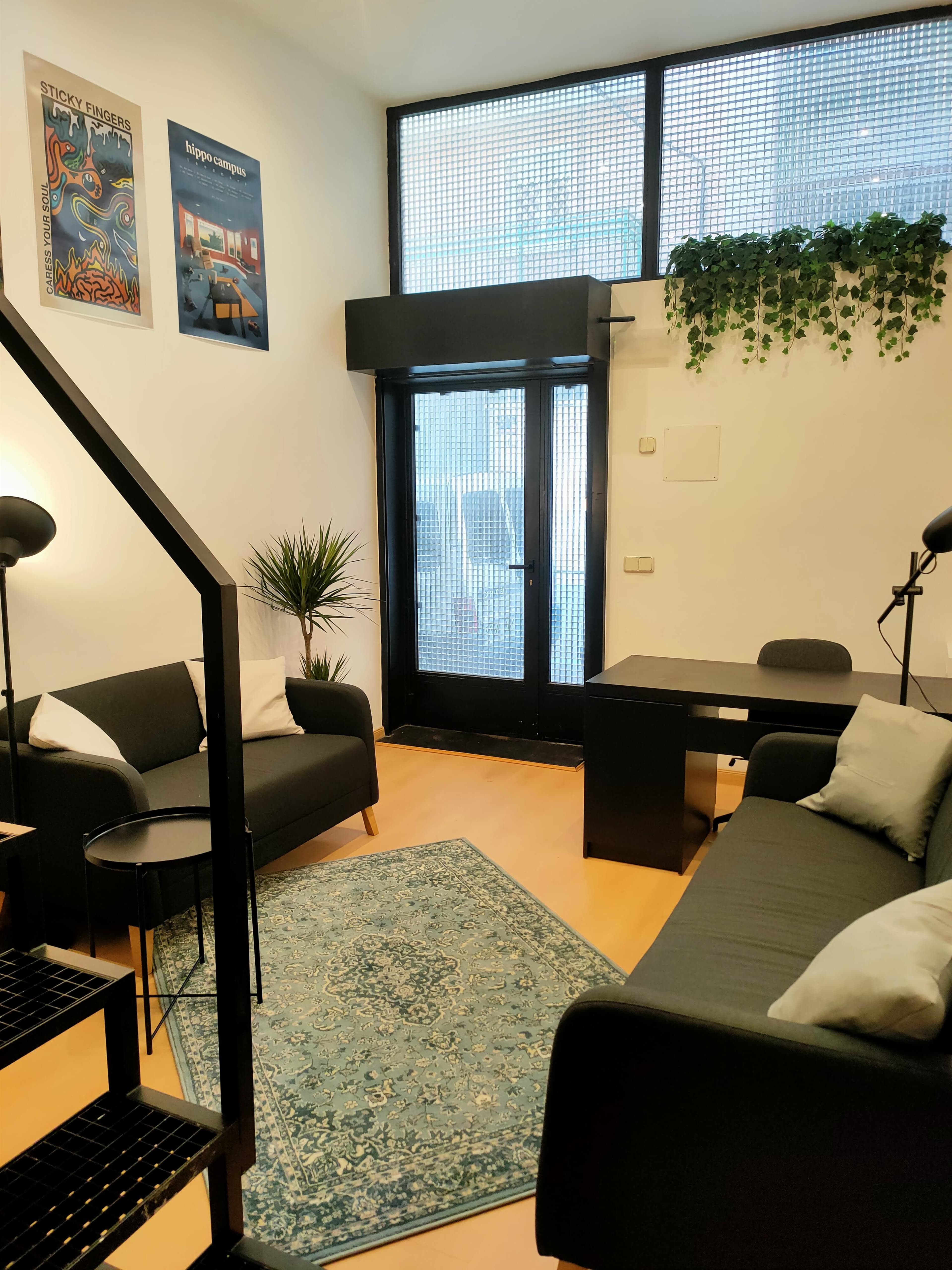 A modern office space with two sofas, a desk, a potted plant, and a decorative area rug, illuminated by natural light through large windows.