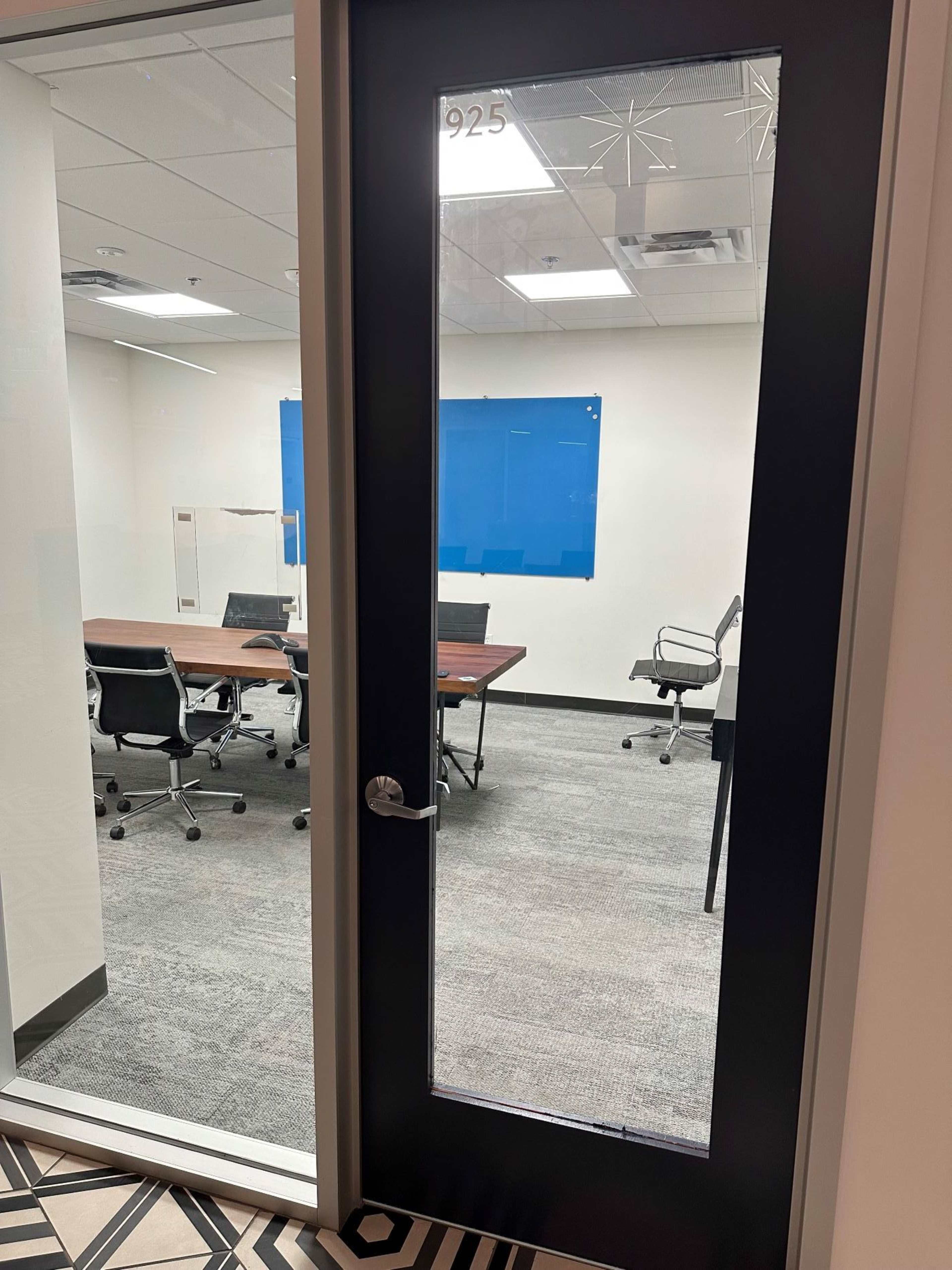 The image shows a glass-paneled door labeled with the number "925" leading into an empty conference room with several chairs and tables inside.