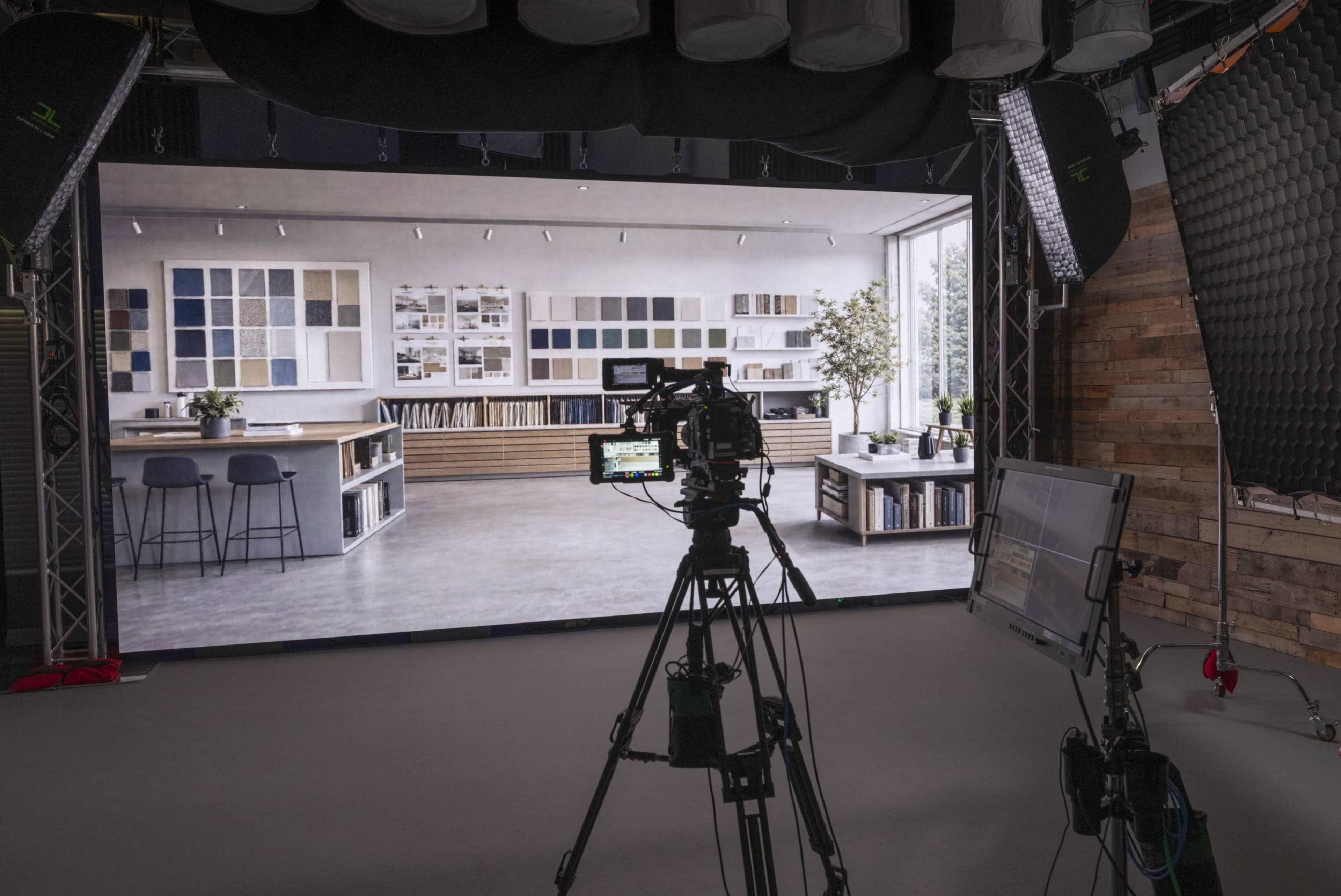 A camera is positioned in front of a large digital backdrop displaying a modern interior design studio with shelves and a large window.