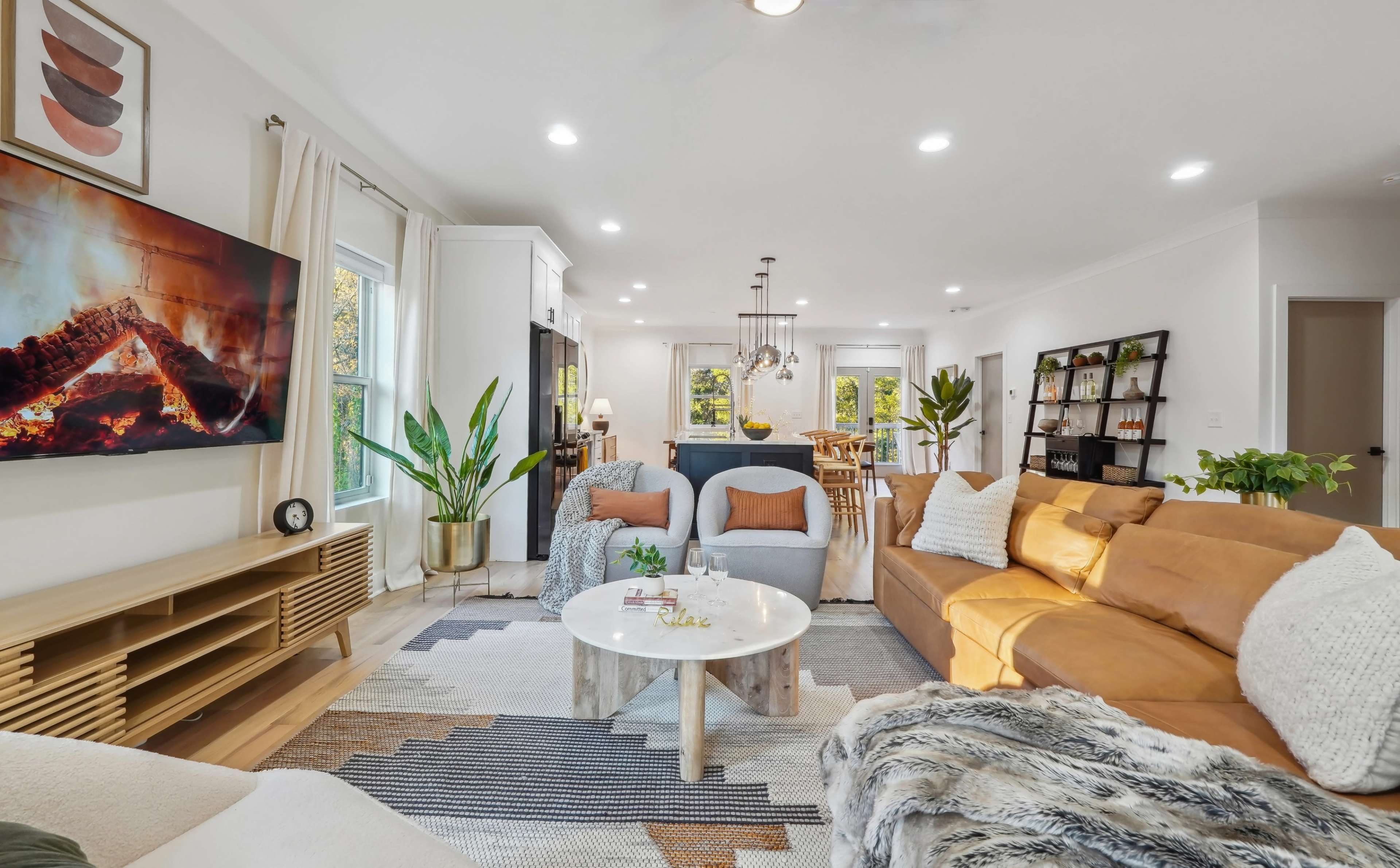 A modern living room features a large sofa, a coffee table, a television mounted on the wall, and various plants, with a dining area visible in the background.