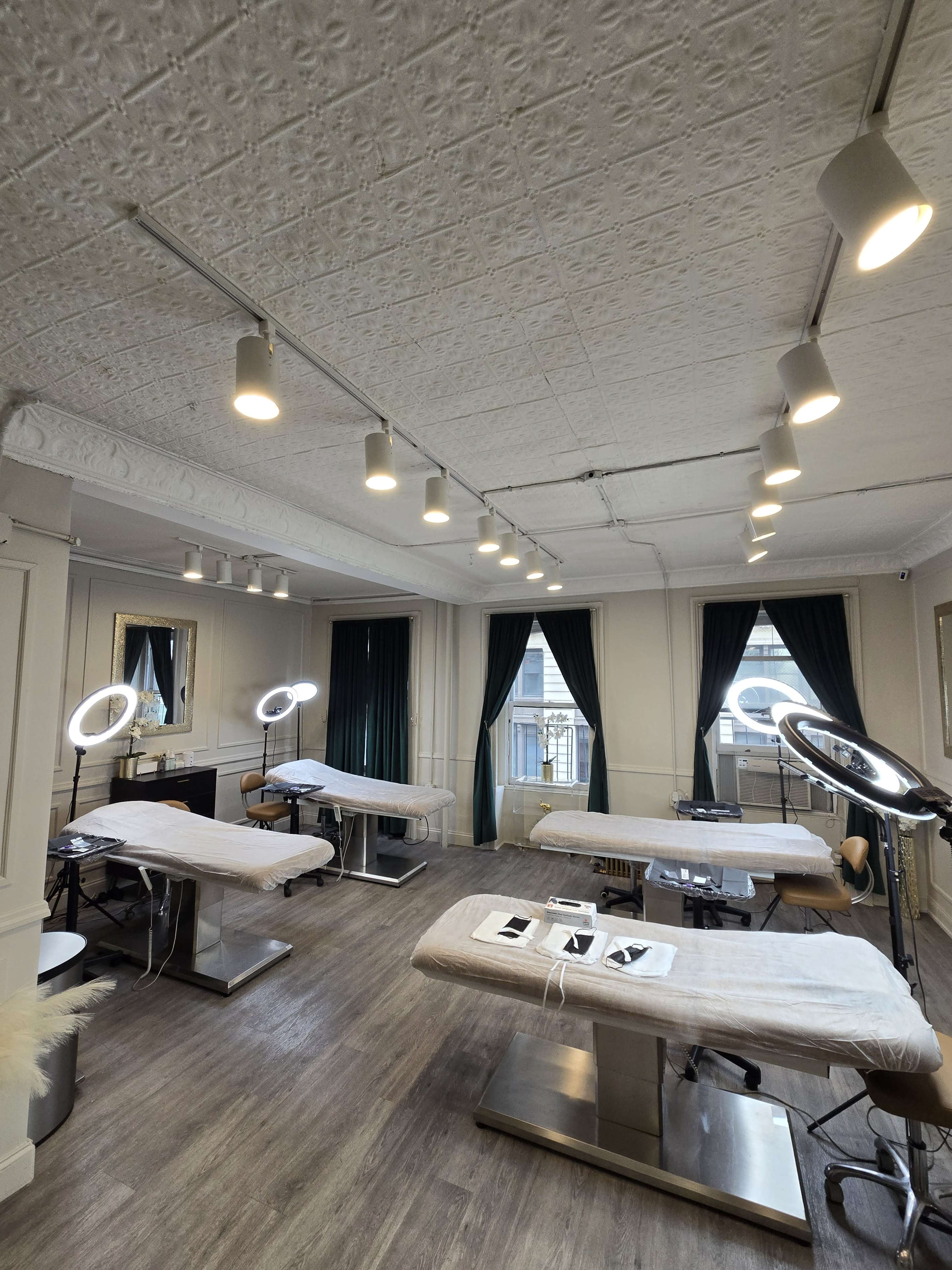 The image shows a well-lit beauty studio with multiple treatment tables, mirrors, and a large window dressed with green curtains.