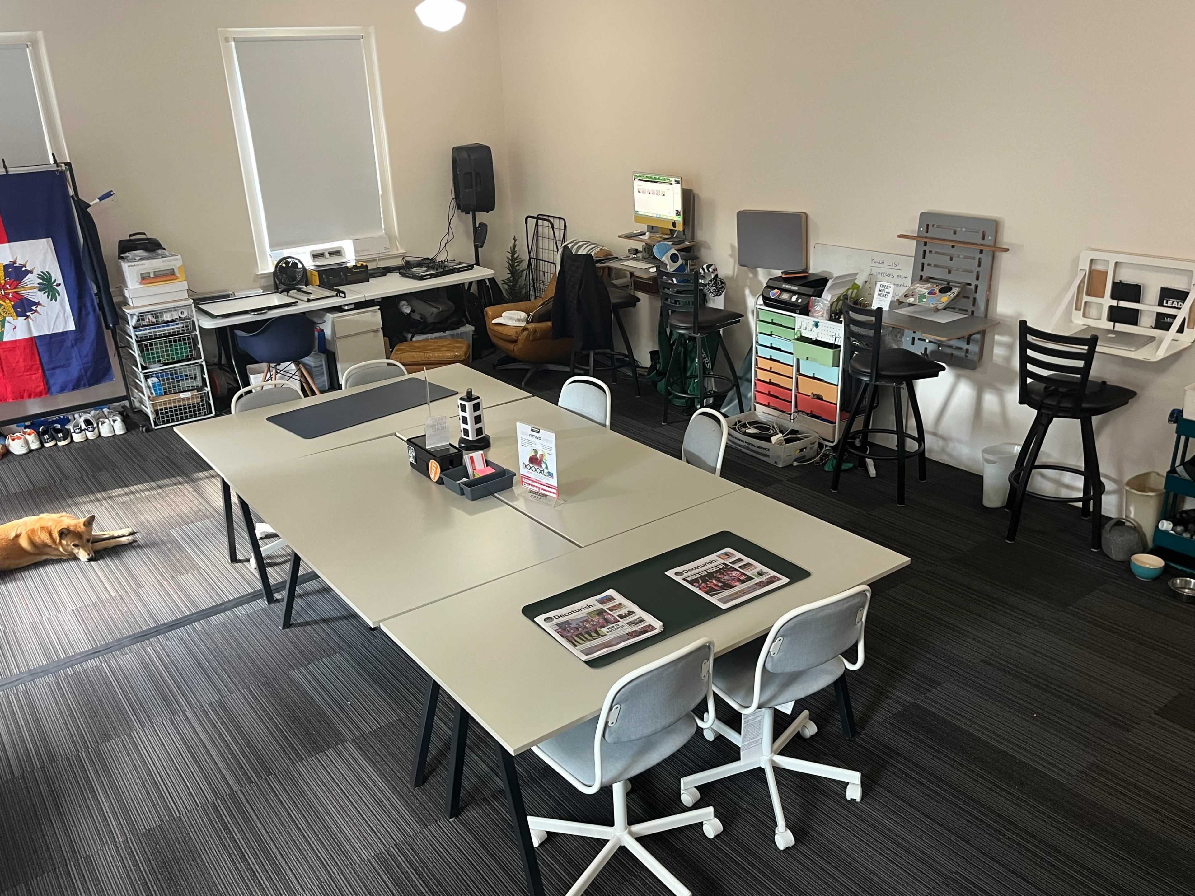 The image shows a spacious, organized workspace featuring a large central table surrounded by chairs, with workstations and equipment along the walls and a dog lying on the floor.