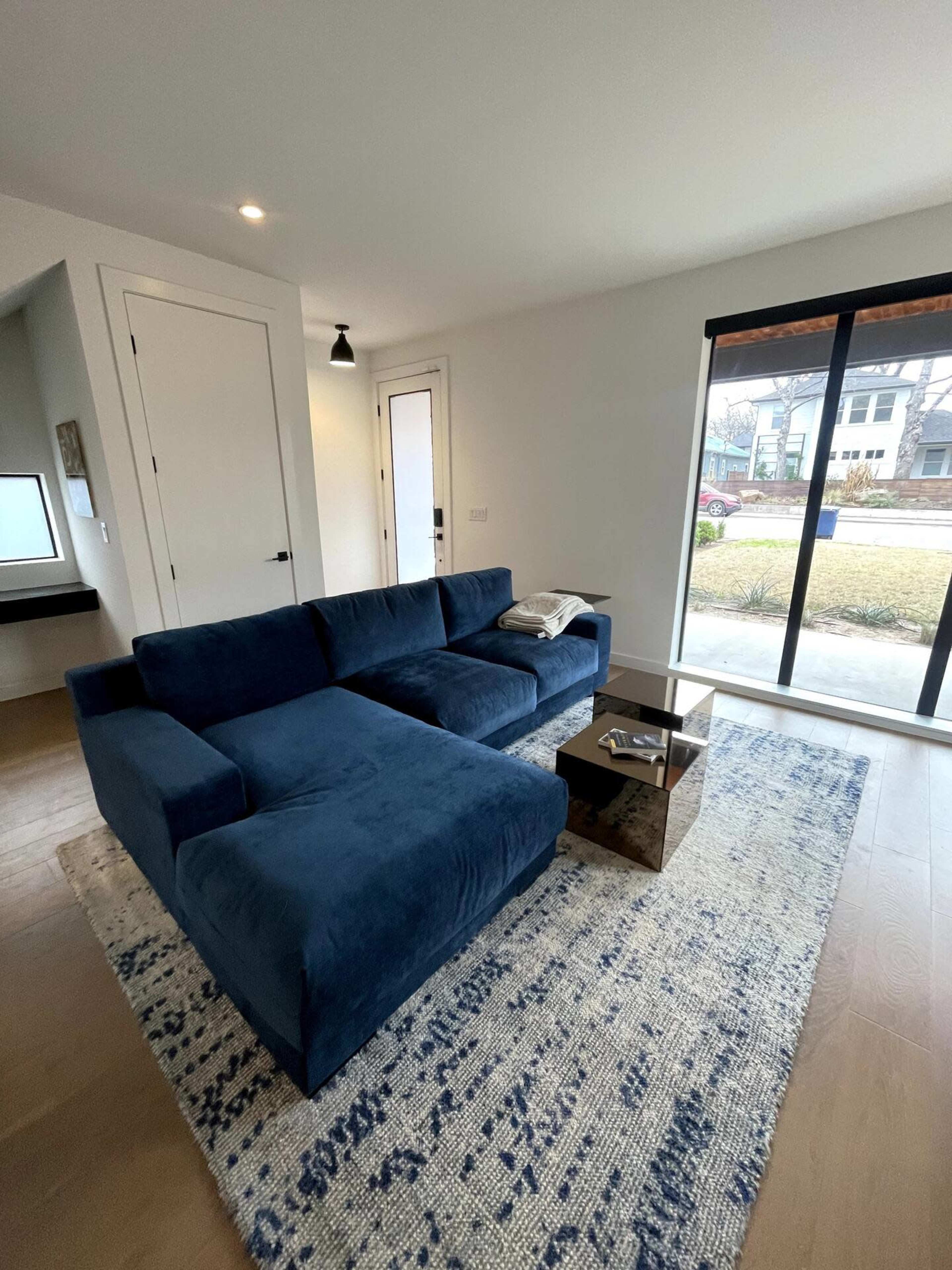 A navy blue sectional sofa is positioned on a patterned rug in a bright living room with large windows and a glass coffee table.