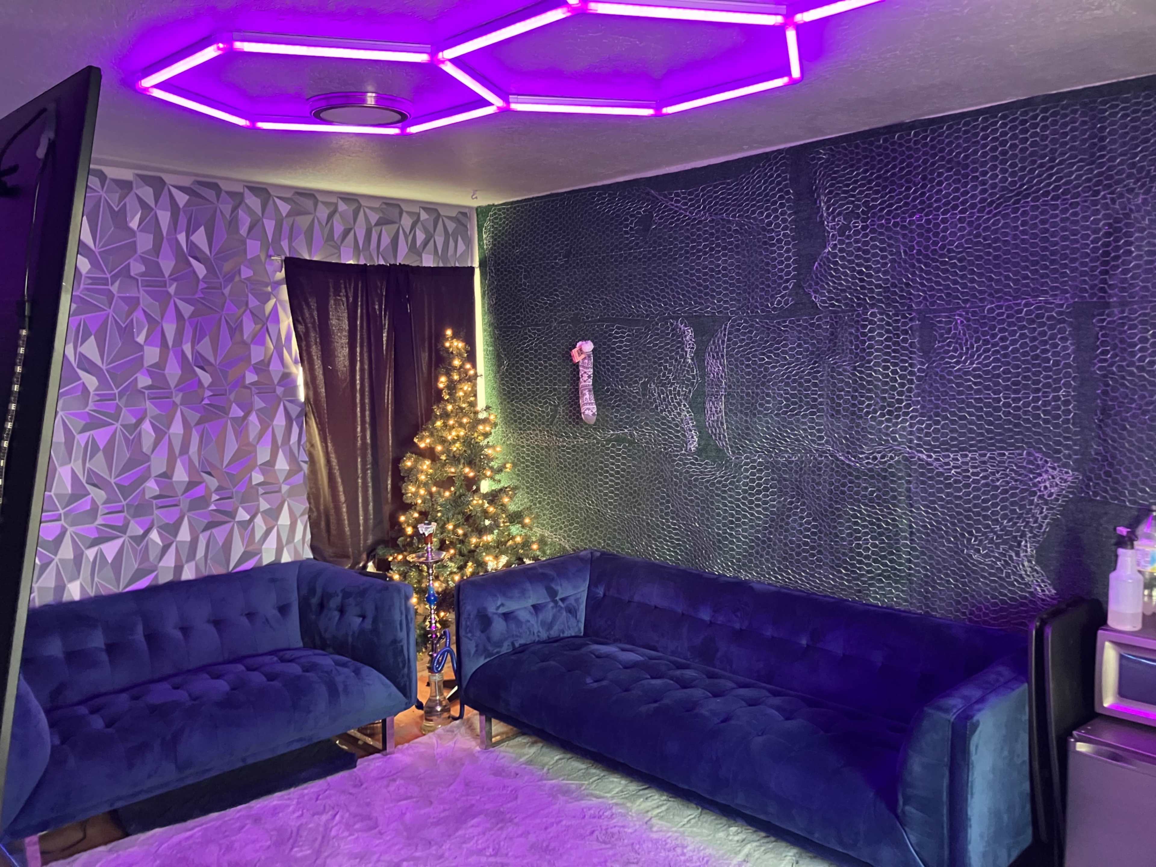 A room features two blue sofas, a small Christmas tree with lights, and a wall with a geometric pattern, illuminated by purple ceiling lights.