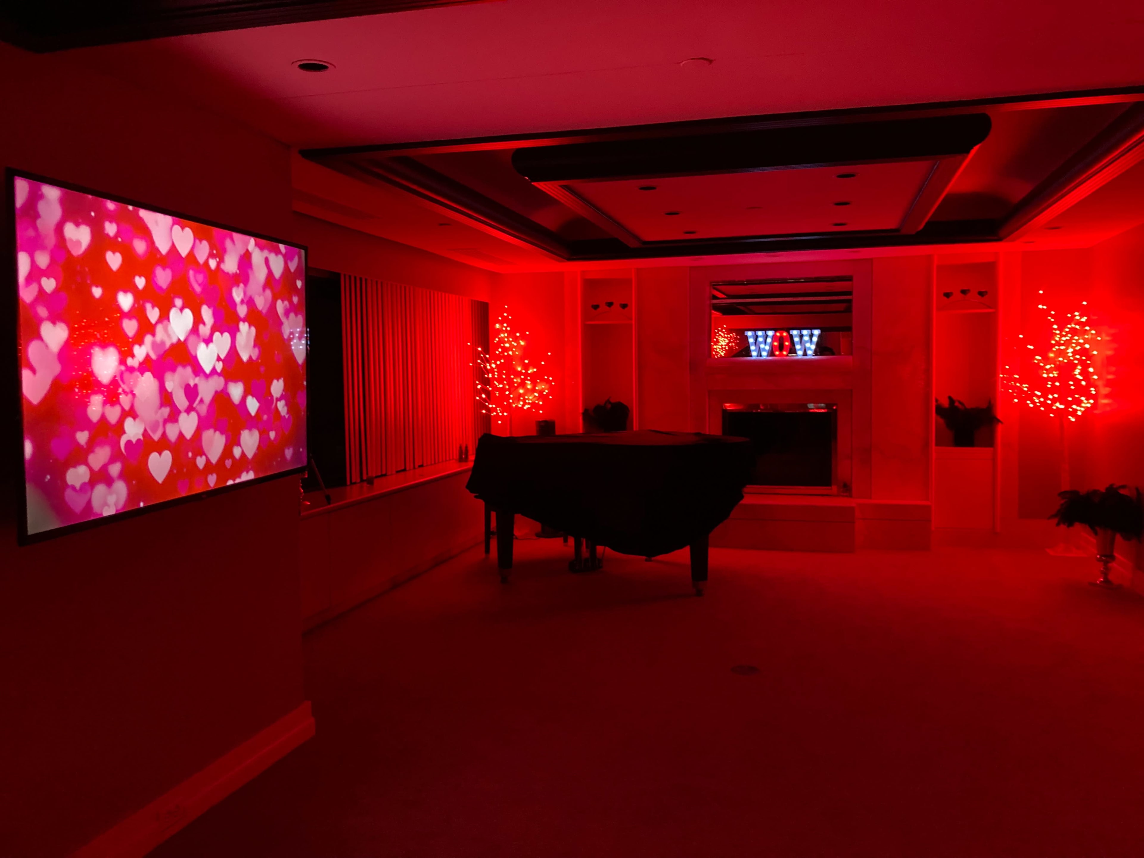 A room is illuminated with red lighting, featuring a large TV displaying a heart-themed graphic, a black grand piano, and decorative lighted trees.