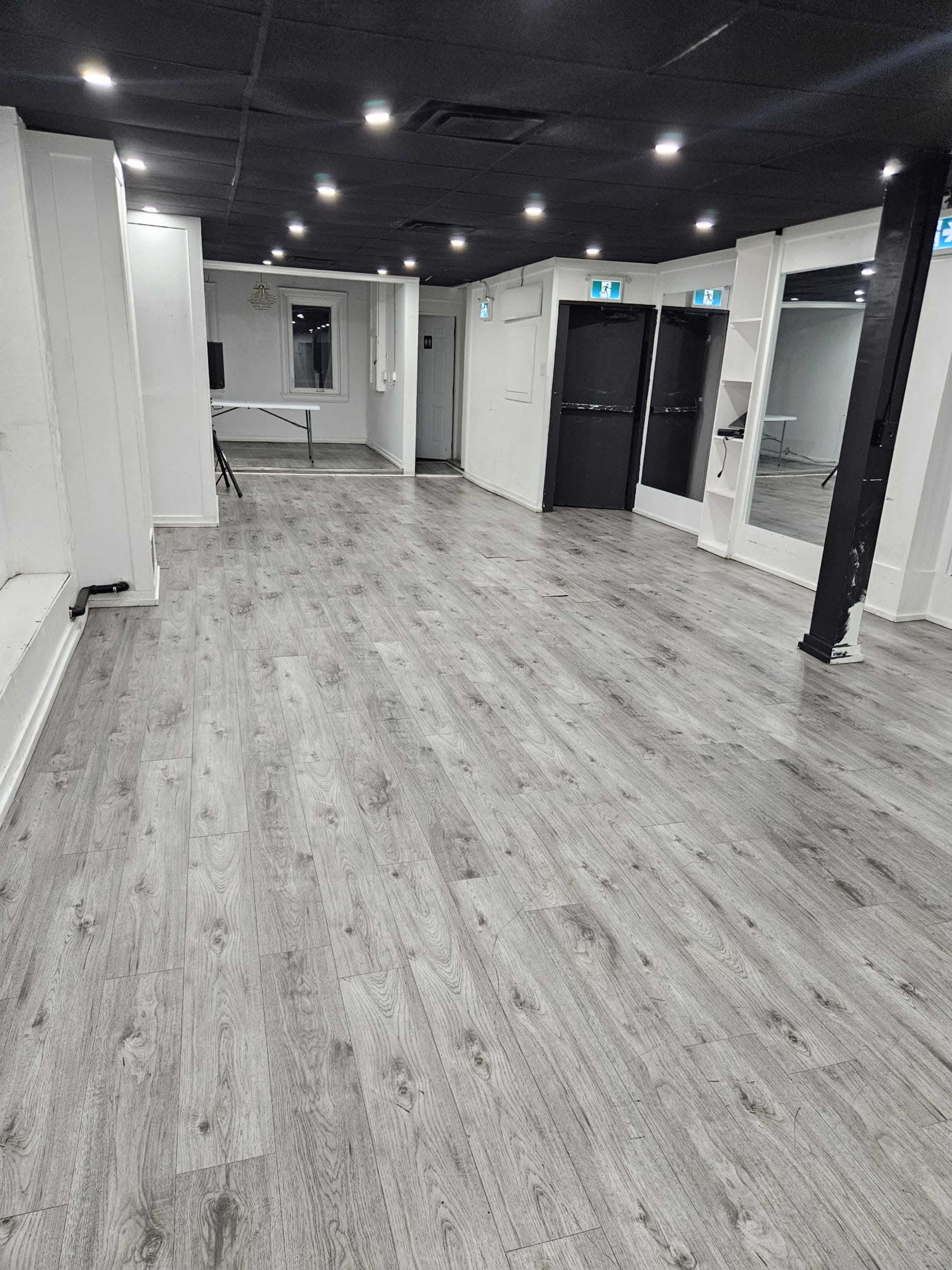 The image shows an empty room with gray wooden flooring, white walls, and several doorways.