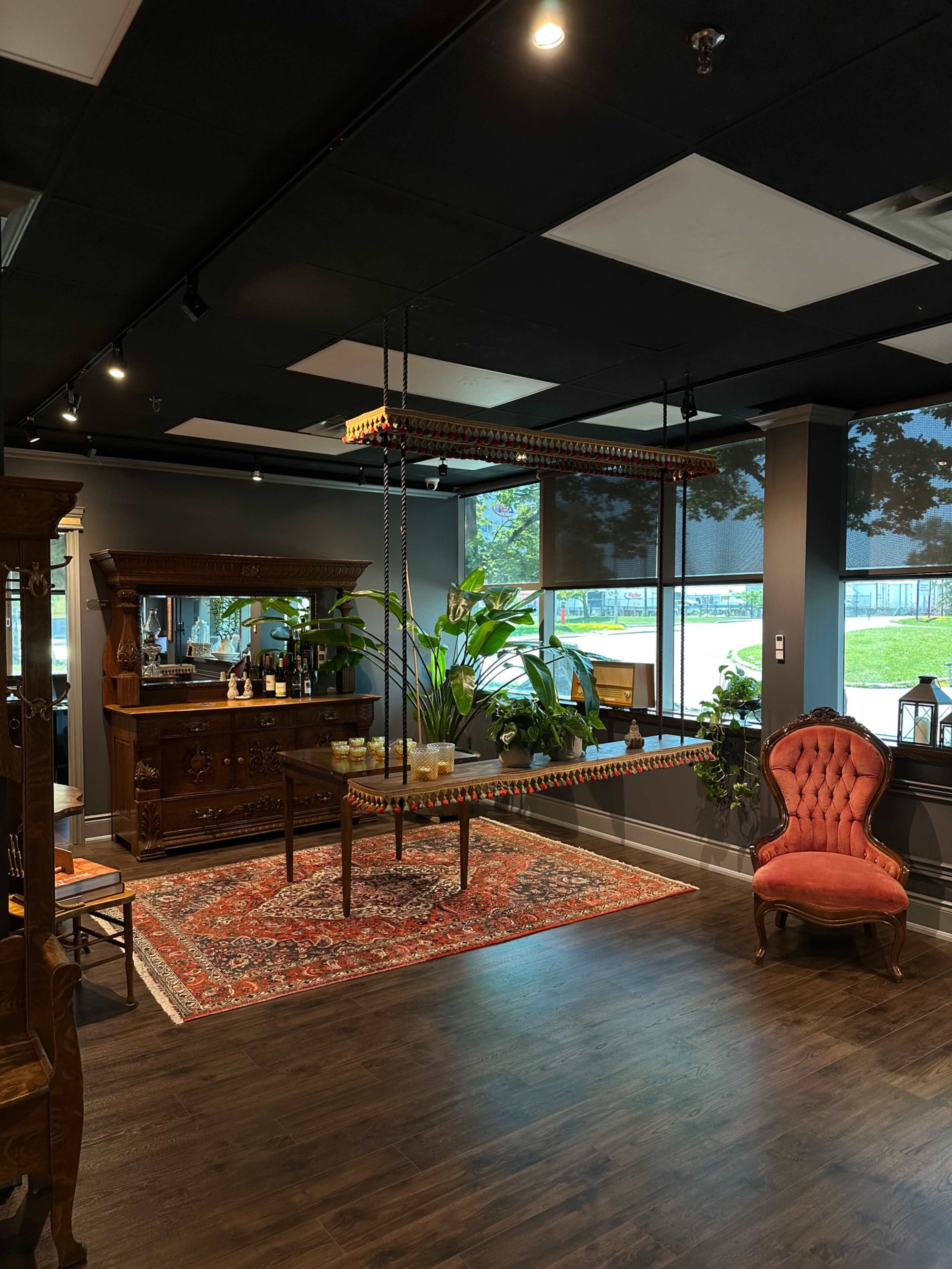 The room features a vintage wooden sideboard, a plush red chair, and a suspended swing above a decorative rug, with large windows allowing natural light to illuminate the space.