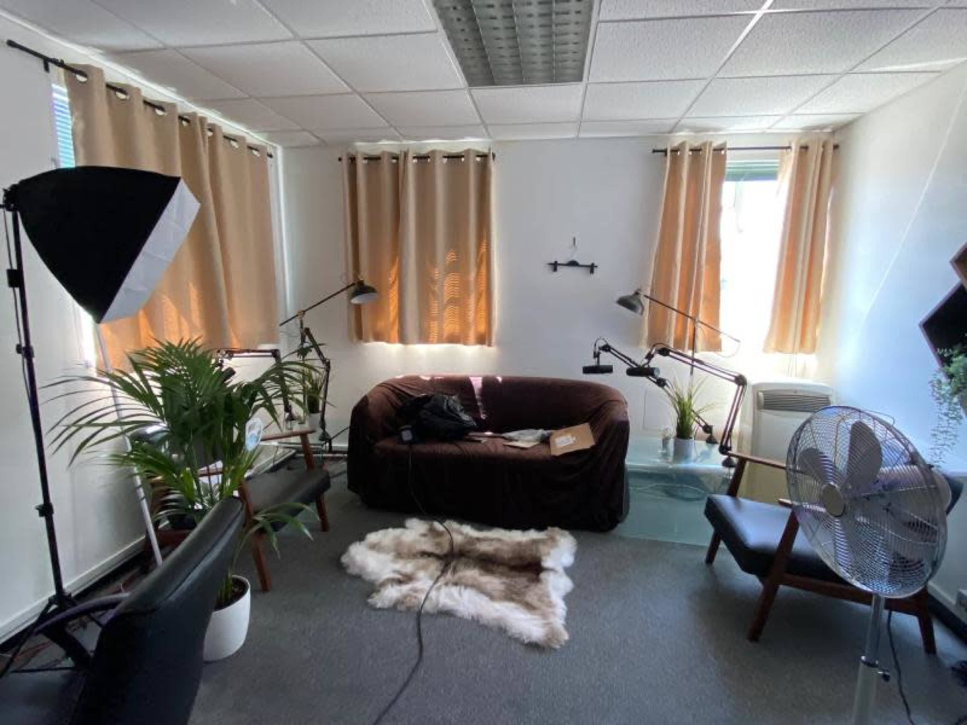 A well-lit office space featuring a brown sofa, two modern chairs, a plant, and various lighting equipment positioned around the room.